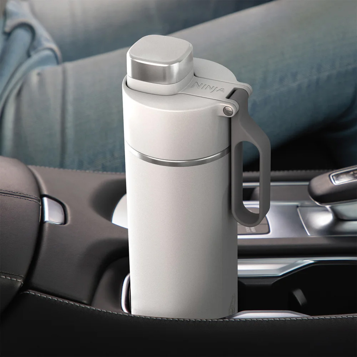 Una bottiglia d'acqua bianca con manico grigio si trova in un portabicchieri per auto. I jeans sono visibili.