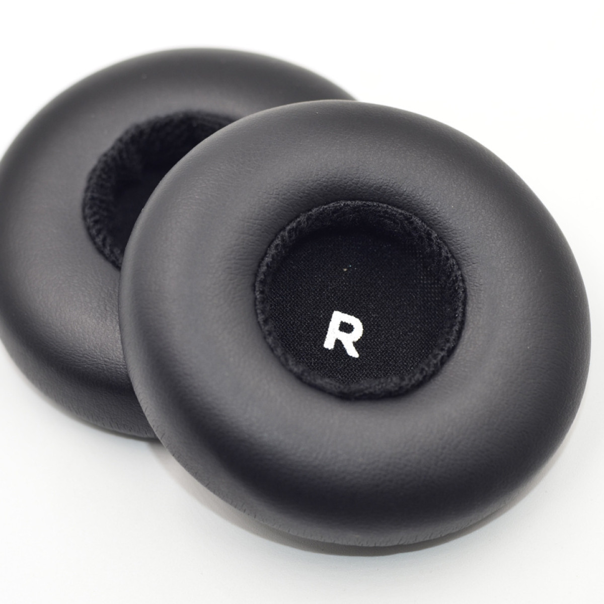 Dwie czarne poduszki nauszne do słuchawek. Jedna ma białe 'R' pośrodku. Są miękkie i okrągłe.