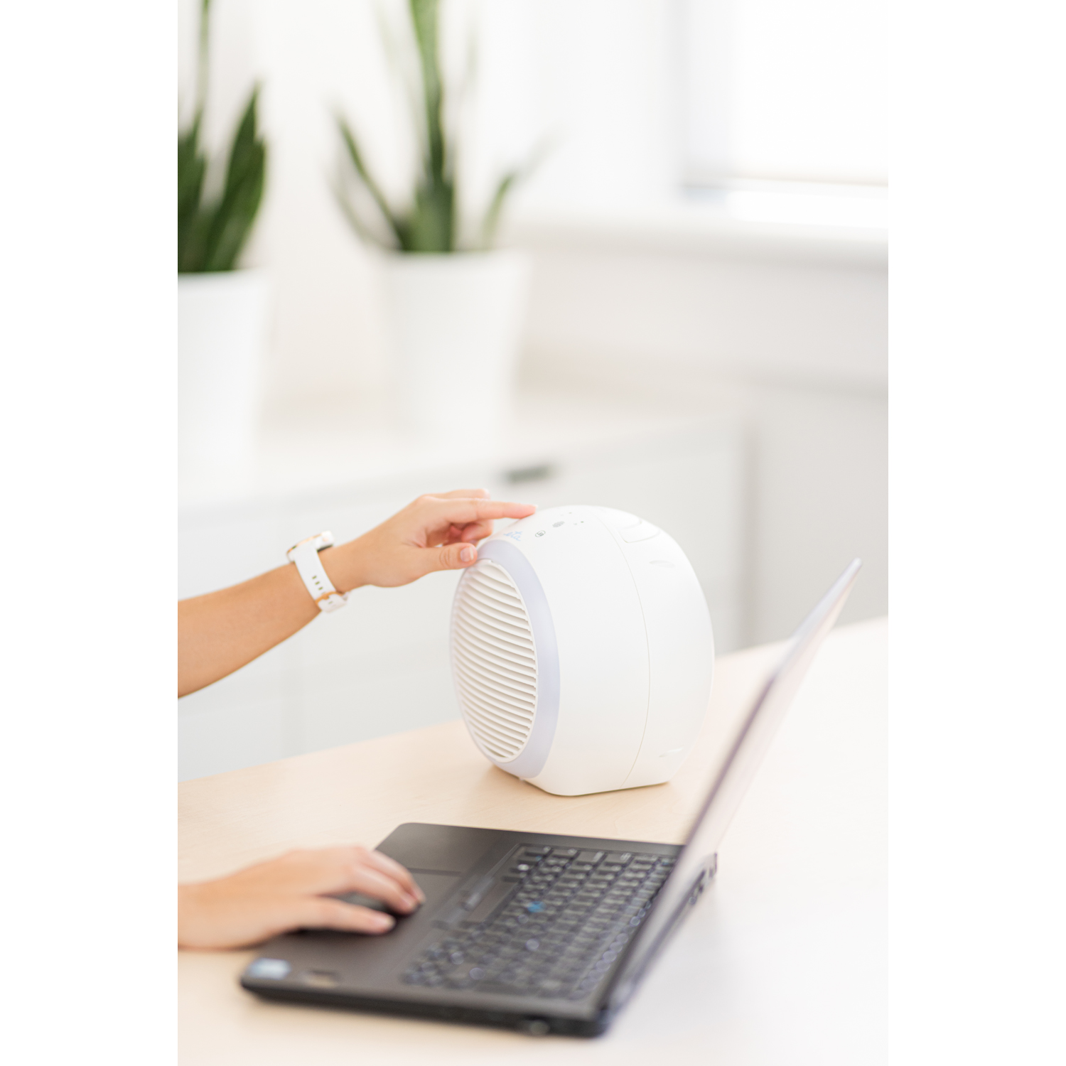 Mano di una persona che regola un ventilatore da scrivania bianco vicino a un laptop su una scrivania in legno chiaro.