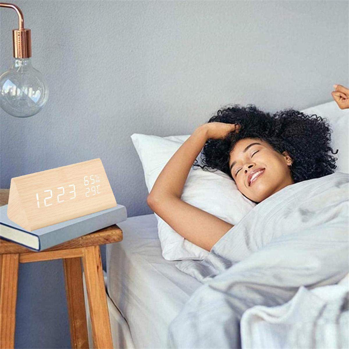Eine Frau im Bett mit einem hölzernen Wecker, der die Uhrzeit und Temperatur anzeigt. Ein Buch liegt neben der Uhr.