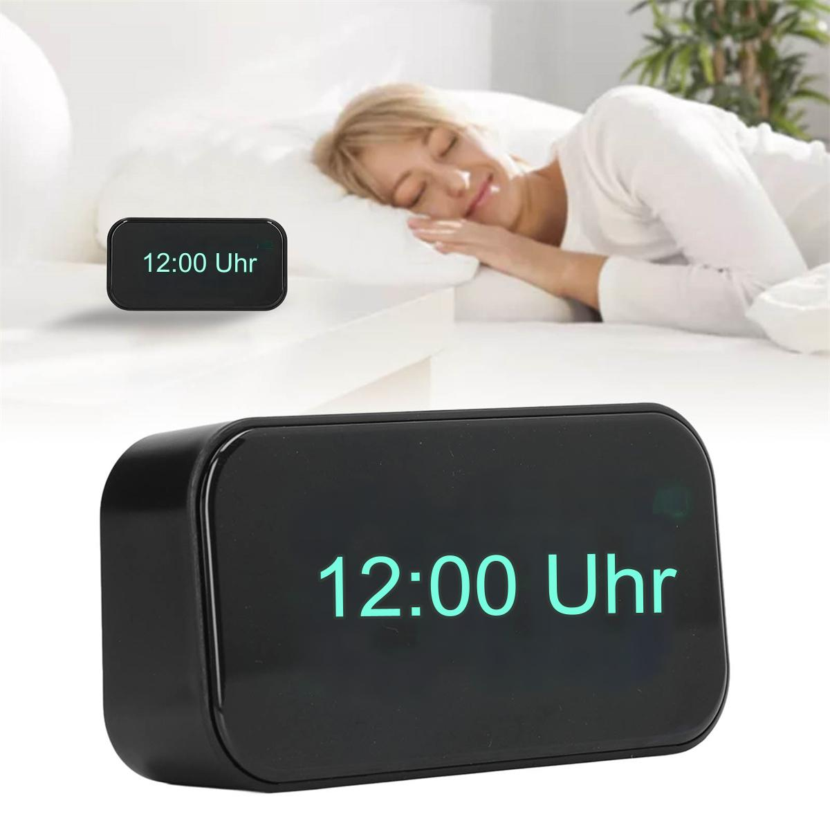 Schwarze Digitaluhr mit '12:00 Uhr'. Im Hintergrund schläft eine Frau.