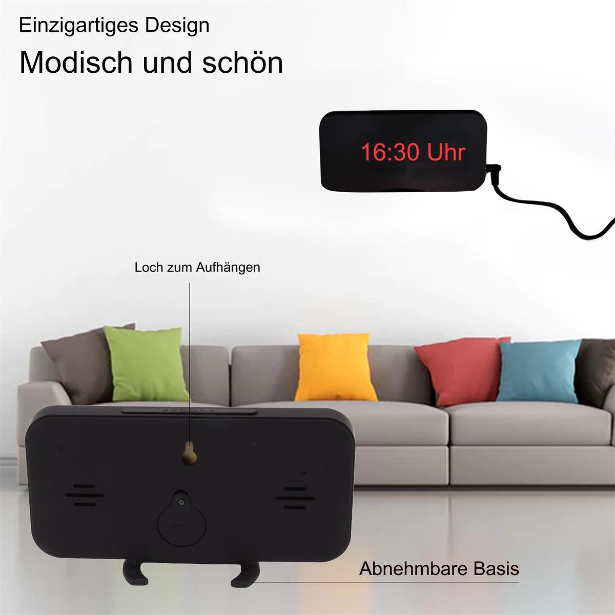 Ein schwarzer Wecker, der 16:30 Uhr anzeigt. Die Uhr hängt an einer weißen Wand, im Hintergrund ein Sofa.