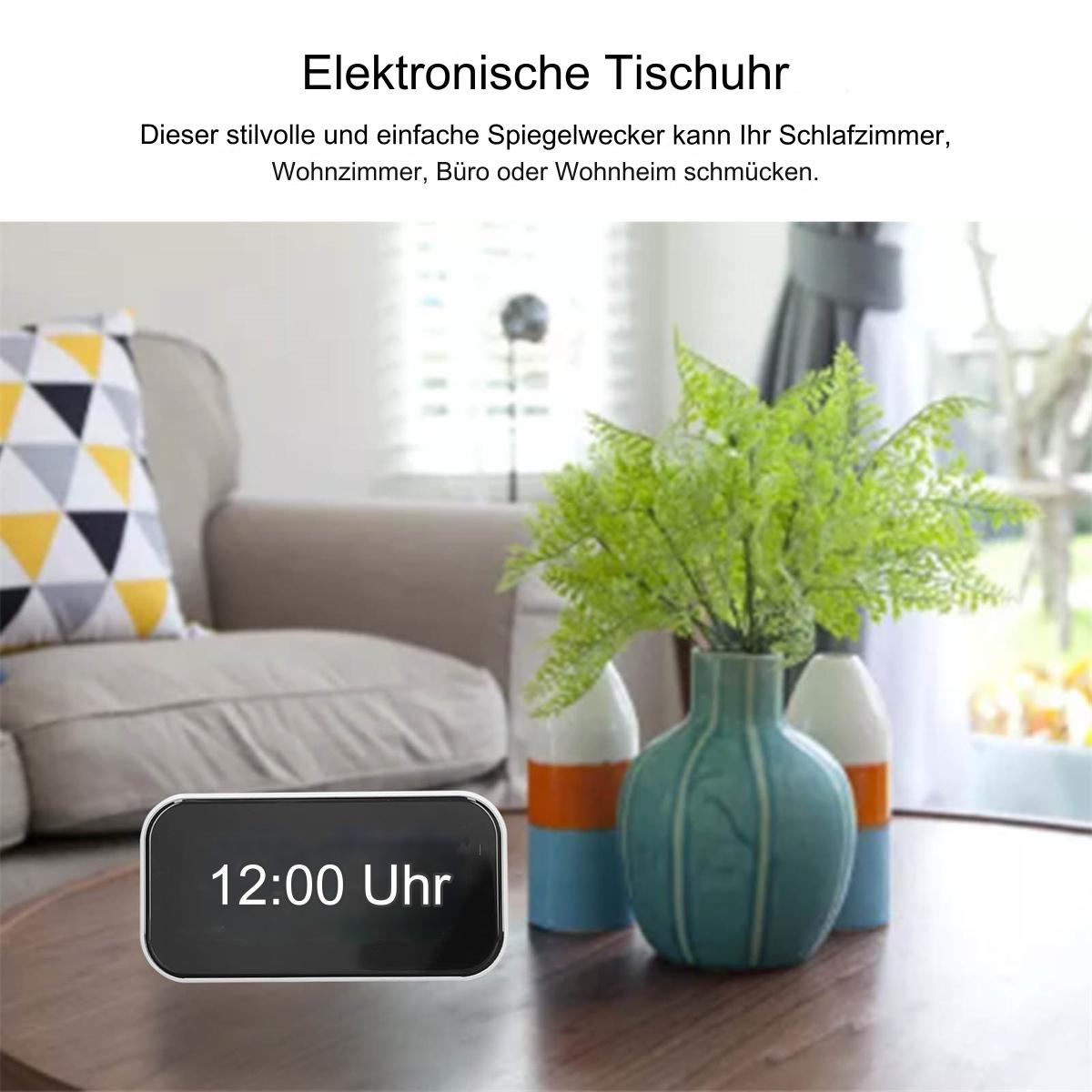 Eine Digitaluhr zeigt 12:00 Uhr an. Sie steht auf einem Tisch mit einer Vase mit grüner Pflanze, in einem Raum mit Sofa.