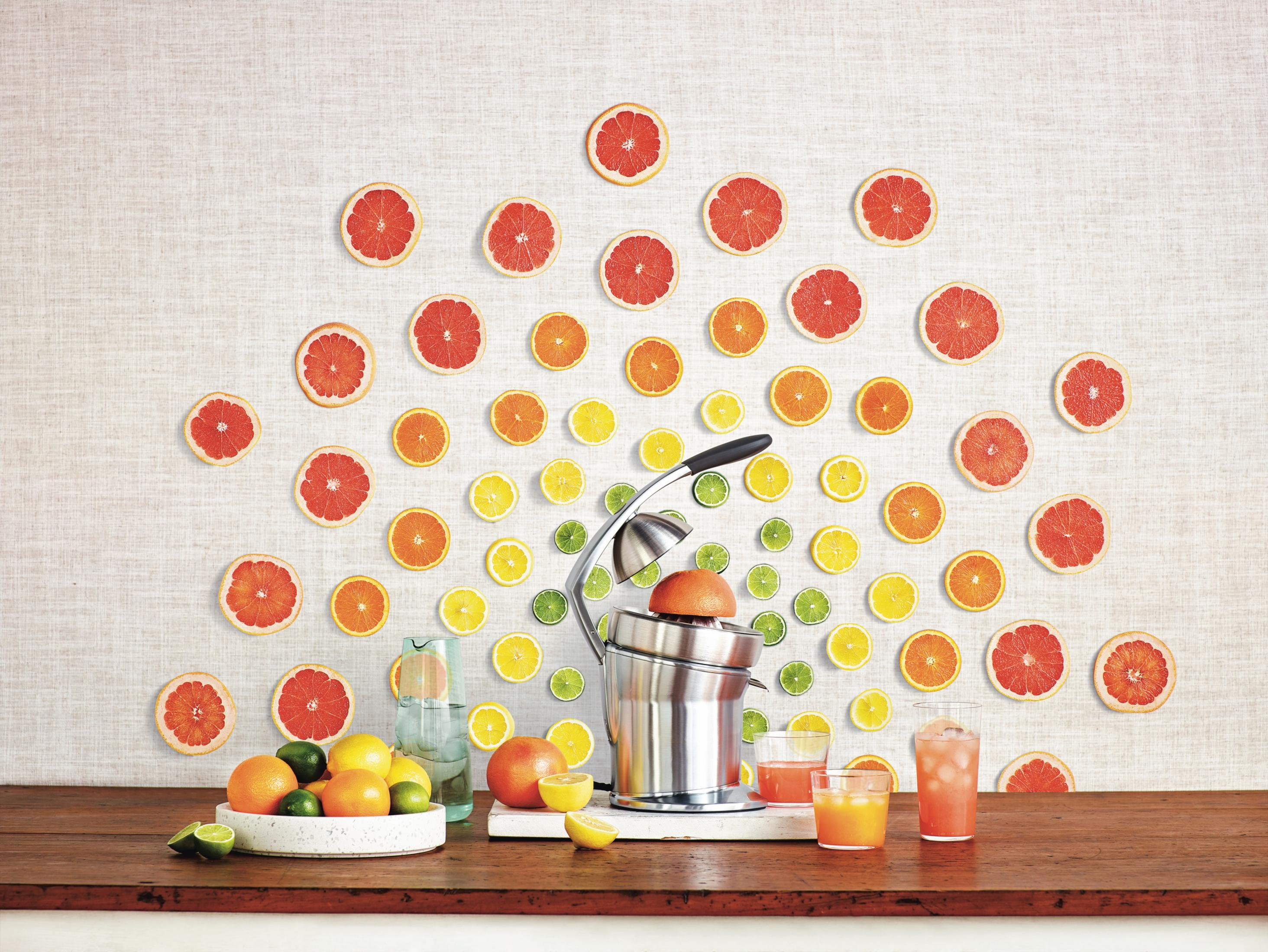Stół z owocami cytrusowymi, wyciskarką do soku i szklankami soku, ułożony pod ścianą.