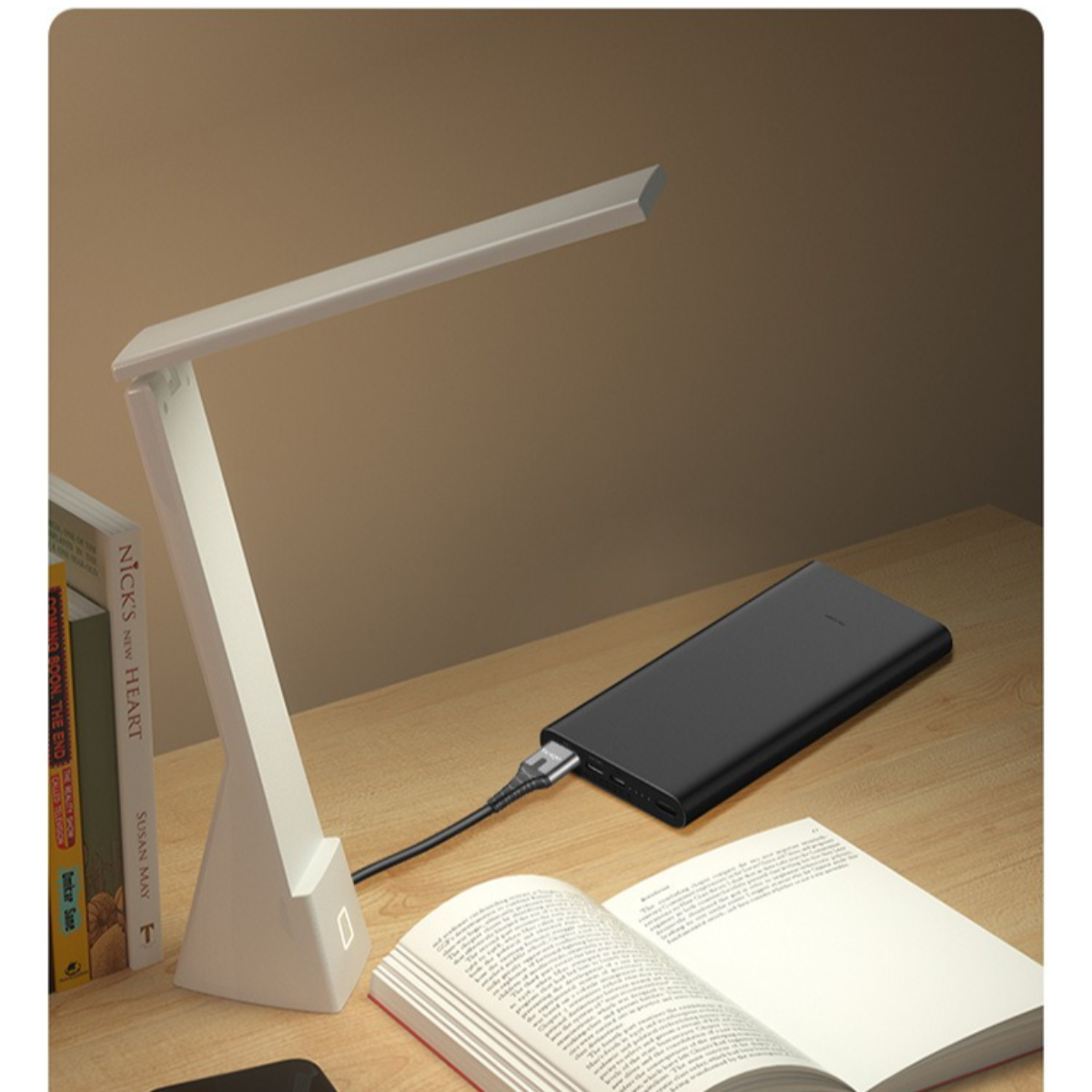 Eine weiße Schreibtischlampe ist an. Eine Powerbank ist mit der Lampe verbunden, mit einem offenen Buch und anderen Büchern.