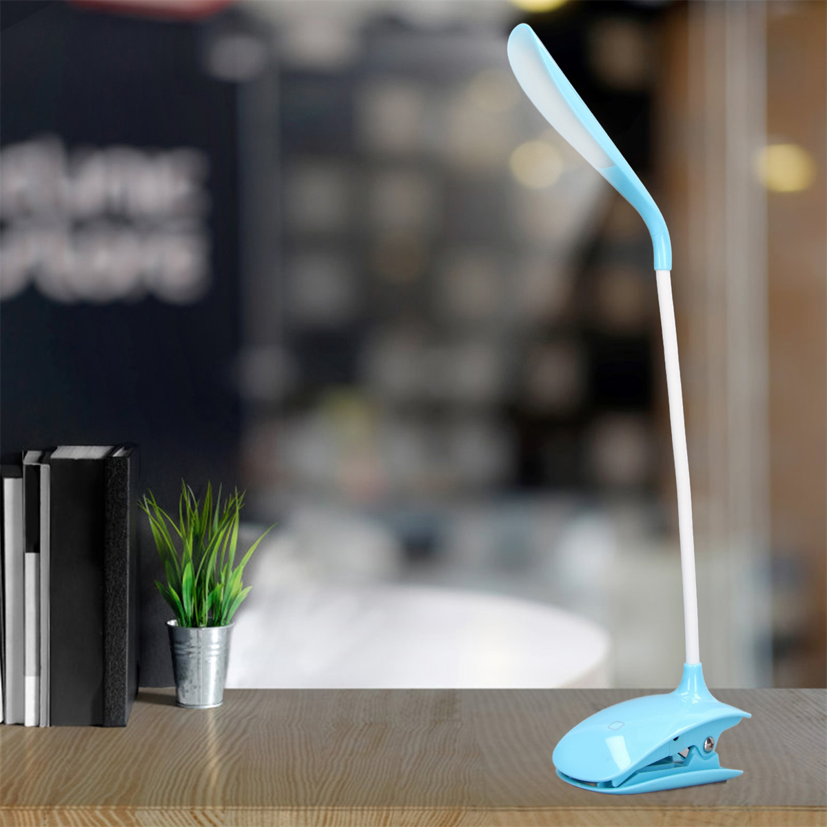 Blaue und weiße Schreibtischlampe mit Clip-Basis. Grüne Topfpflanze und Bücher auf einem Holzschreibtisch. Unscharfer Hintergrund.