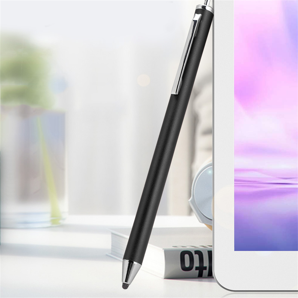 Schwarzer Eingabestift lehnt an einem weißen Tablet. Ein Buch liegt auf dem Tisch. Fenster im Hintergrund.