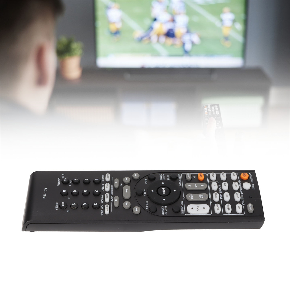 Eine schwarze Fernbedienung ist im Vordergrund. Im Hintergrund schaut eine Person fern. Der Fernseher zeigt ein Fußballspiel.
