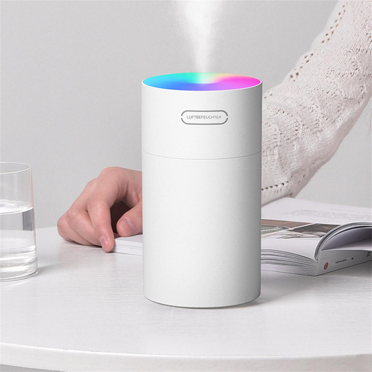 Ein weißer Luftbefeuchter mit farbigem Licht oben, eine Hand und ein Glas Wasser stehen auf einem Tisch.