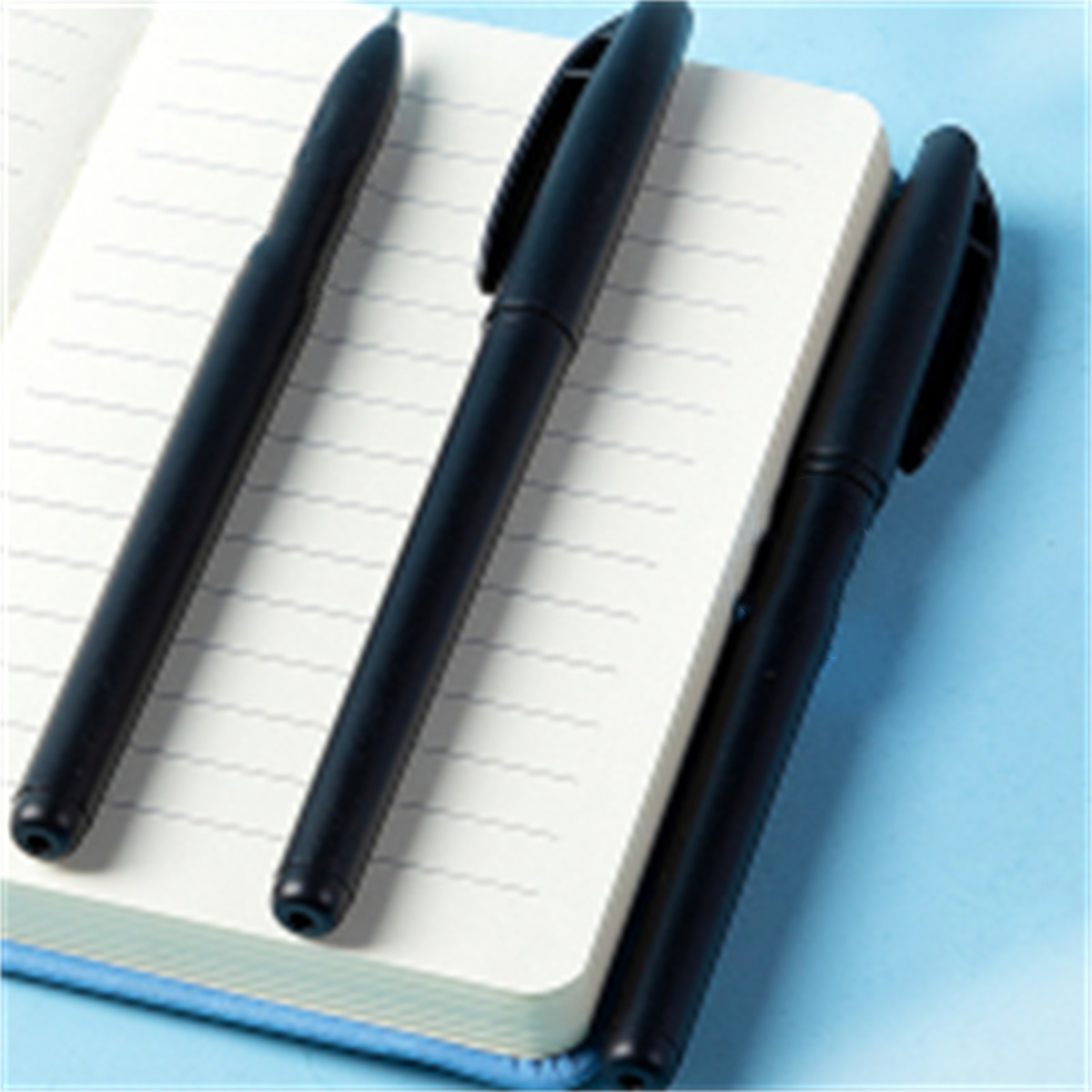 Drei schwarze Stifte auf einem weißen Notizbuch auf blauem Untergrund.