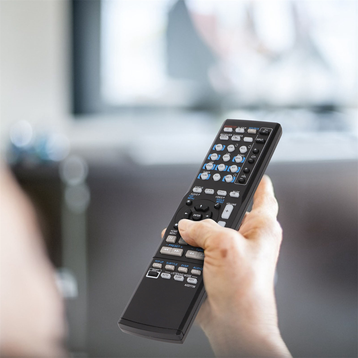 Eine Hand, die eine schwarze Fernbedienung mit vielen Knöpfen hält, mit einem verschwommenen Fernseher im Hintergrund.