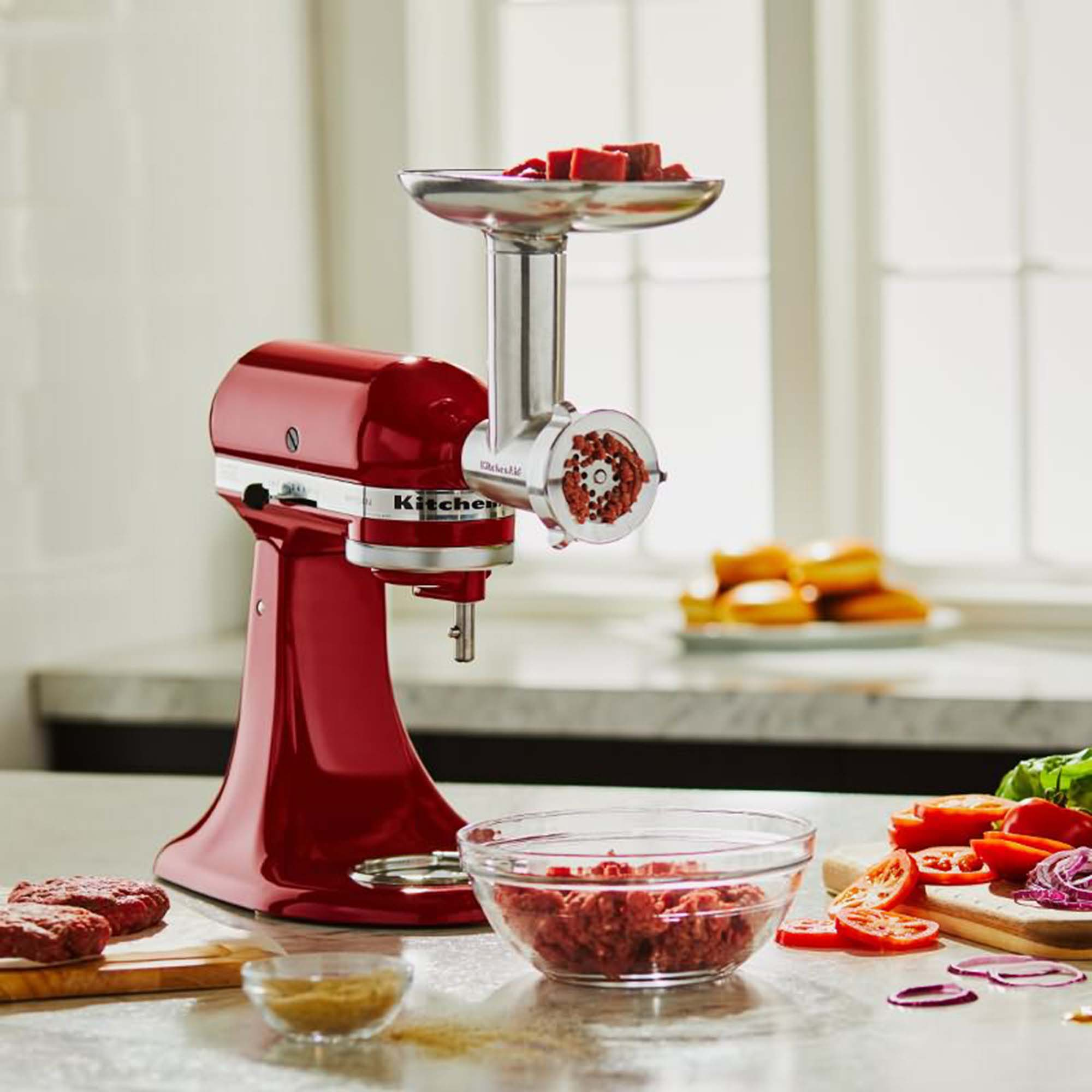Rode KitchenAid mixer met vleesmolen opzetstuk, kom met gehakt en ingrediënten. Wit oppervlak.