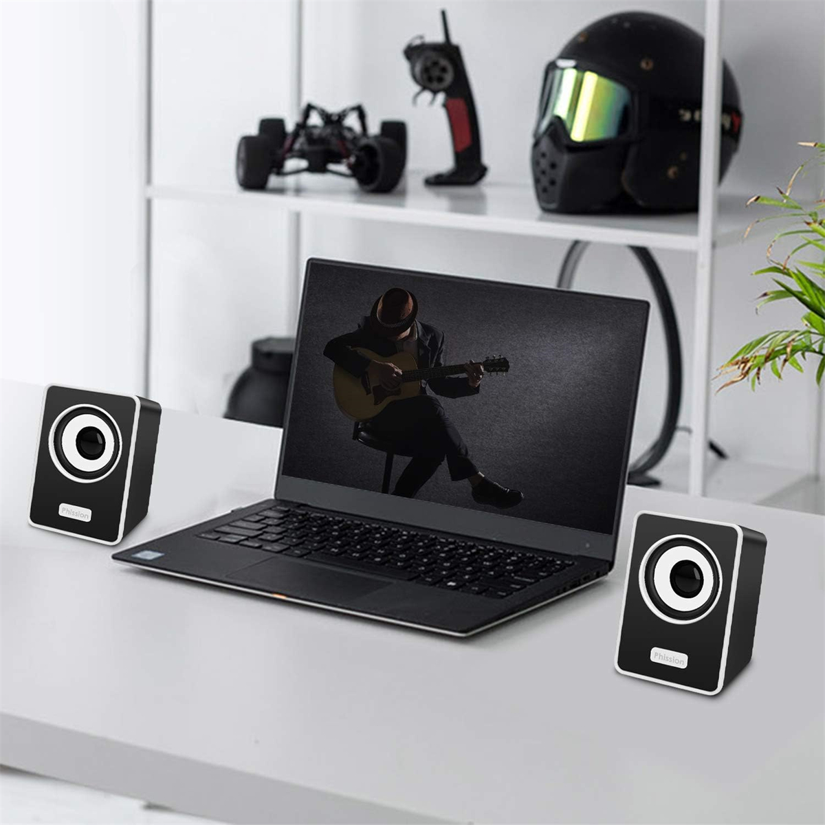 Zwei schwarze Lautsprecher und ein Laptop auf einem weißen Schreibtisch, mit einem Mann, der Gitarre spielt.