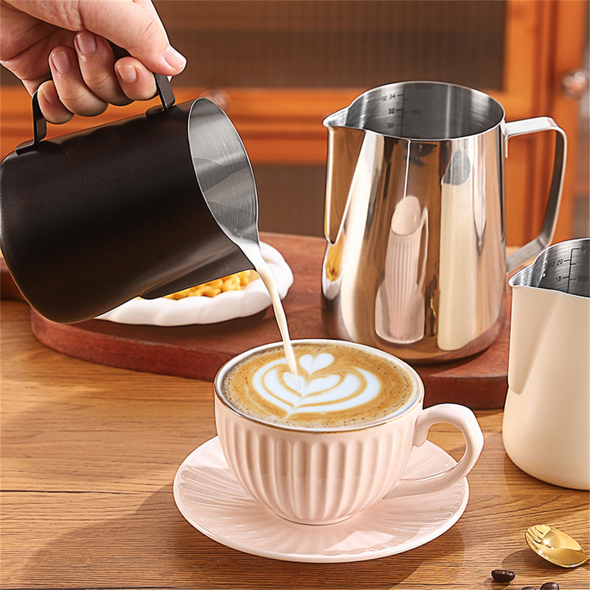 Milch in Kaffee mit Latte Art gießen, mehrere Metallkannen im Hintergrund.