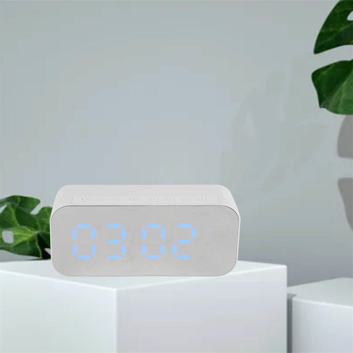 Eine weiße Digitaluhr zeigt die Uhrzeit an, aufgestellt auf einer weißen Plattform mit grünen Pflanzen im Hintergrund.