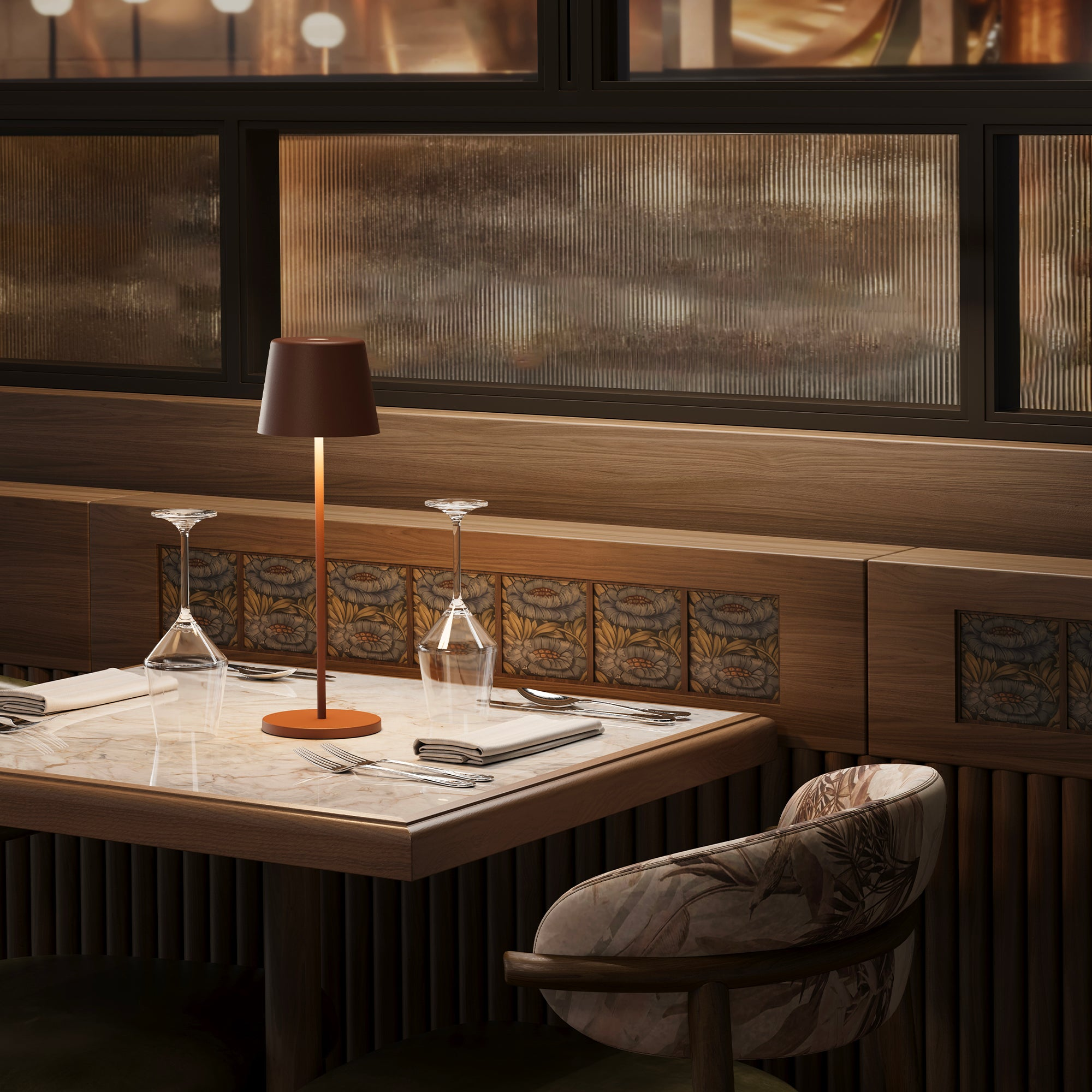 Eine Lampe mit braunem Schirm und langem, dünnem, orangefarbenem Ständer. Sie steht auf einem Marmortisch. Ein Restauranthintergrund.