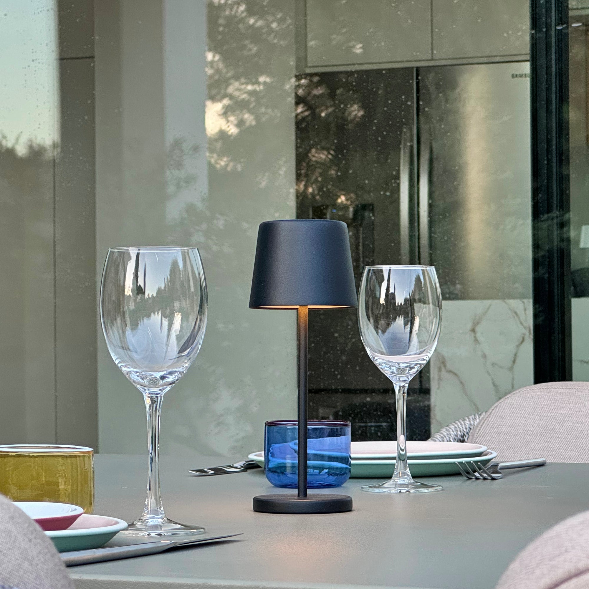 Gedeckter Tisch im Freien mit Weingläsern, einer schwarzen Lampe und einem blauen Glas. Im Hintergrund ein Gebäude.