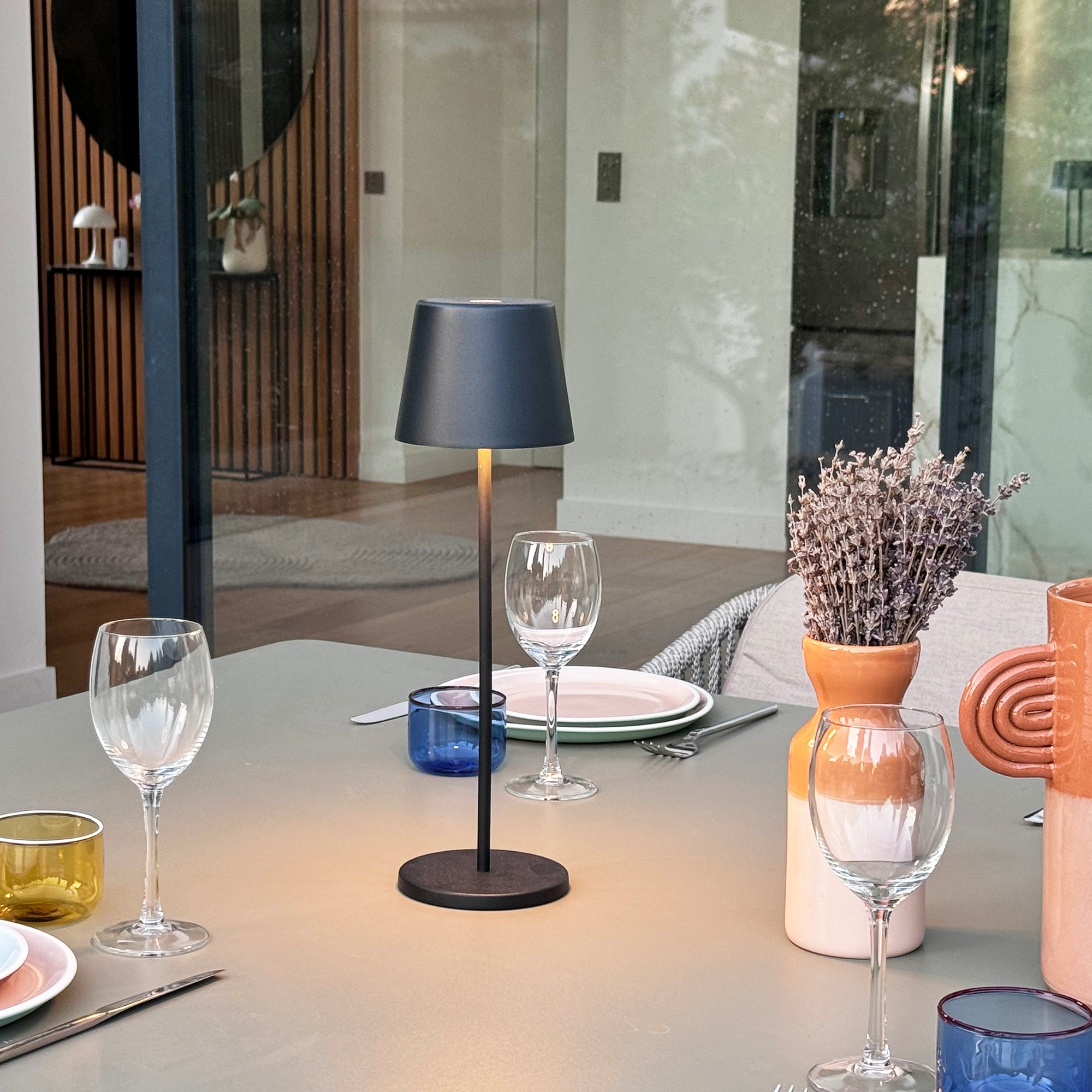 Tisch mit Lampe, Weingläsern und Blumenvase im Freien.