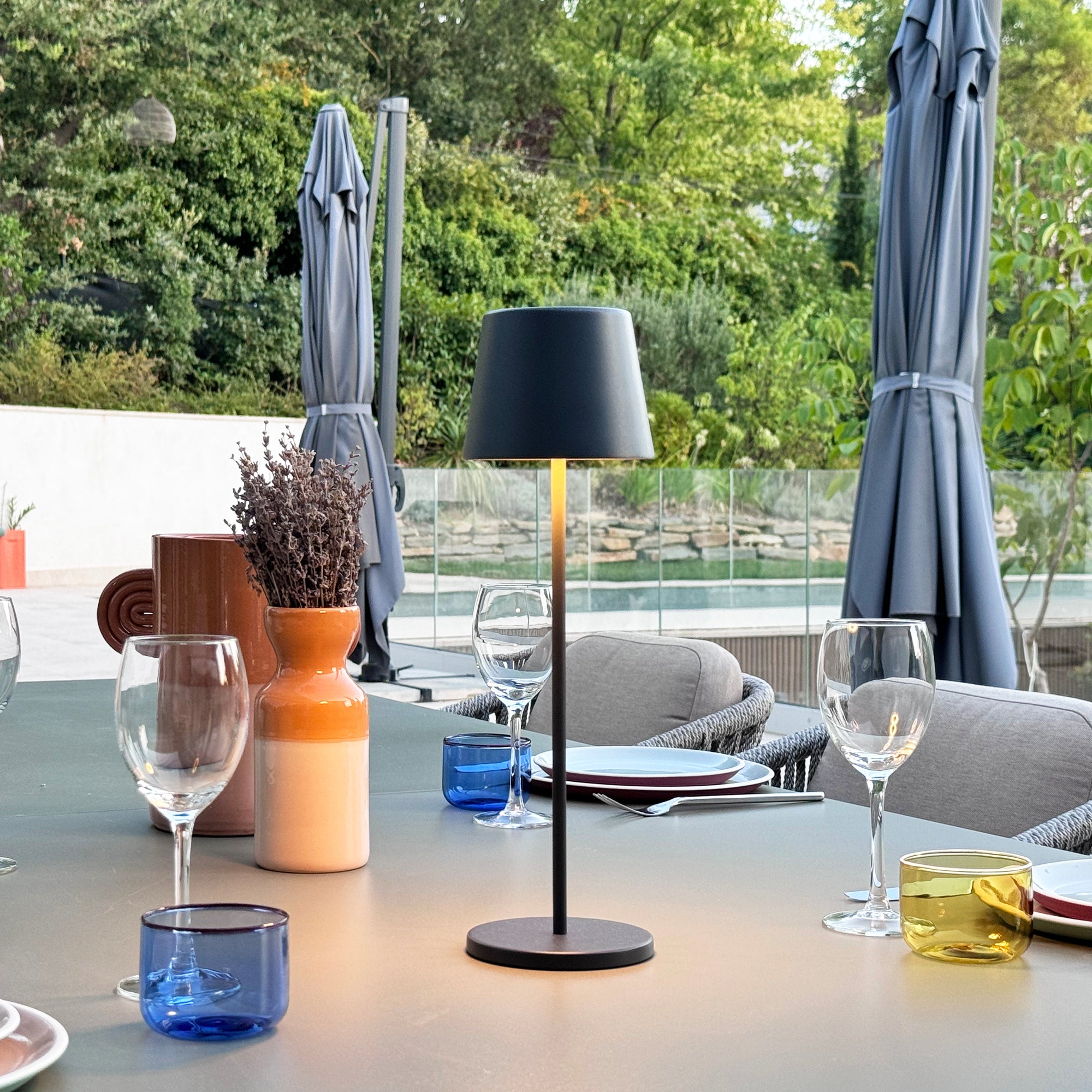 Tisch mit Lampe, Gläsern und Vase. Sonnenschirme, Wasser und Grün im Hintergrund.