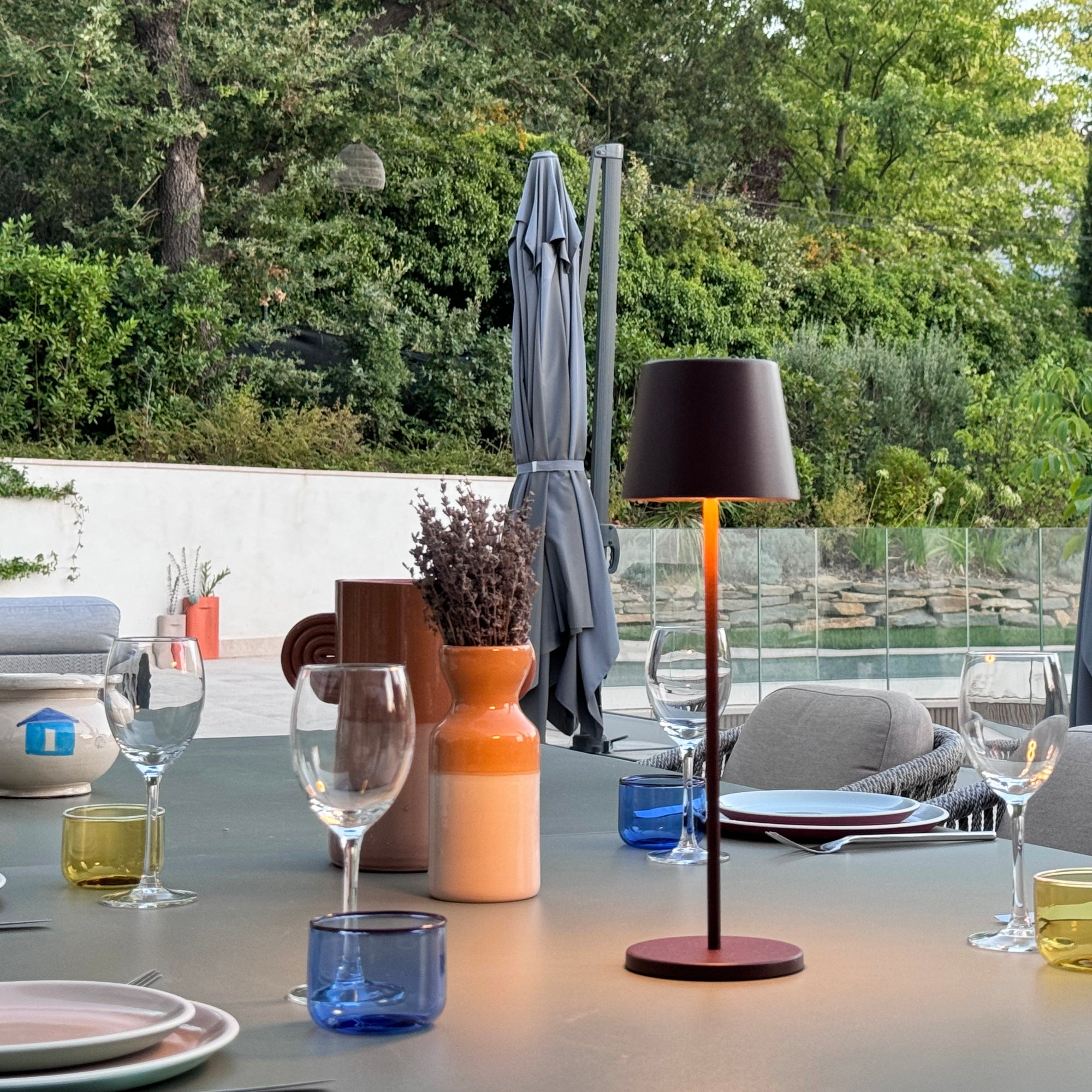Ein Tisch für ein Abendessen im Freien mit Lampe, Weingläsern und anderen dekorativen Gegenständen gedeckt.