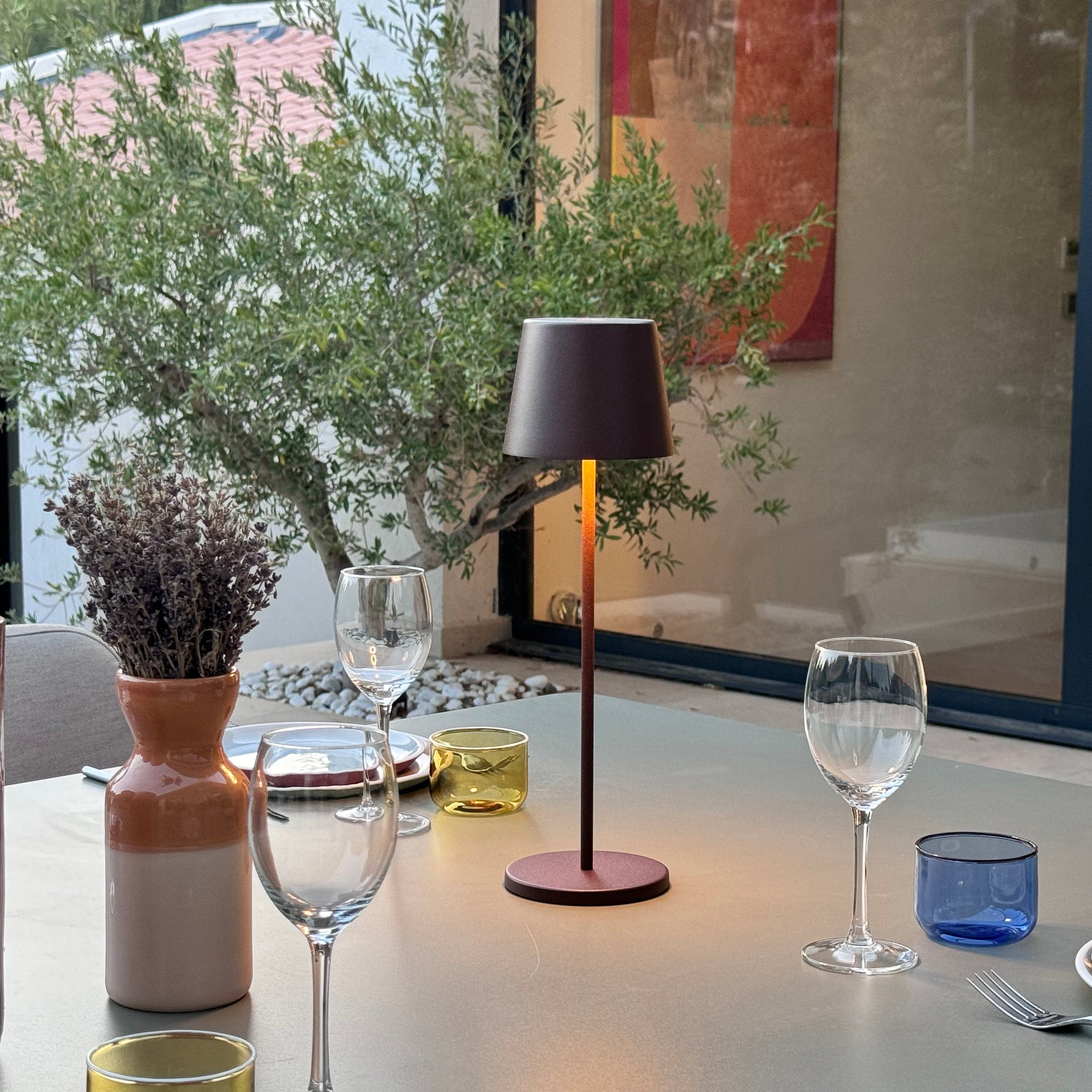 Ein Tisch mit Gläsern, einer Lampe und einer Vase mit Pflanzen draußen.