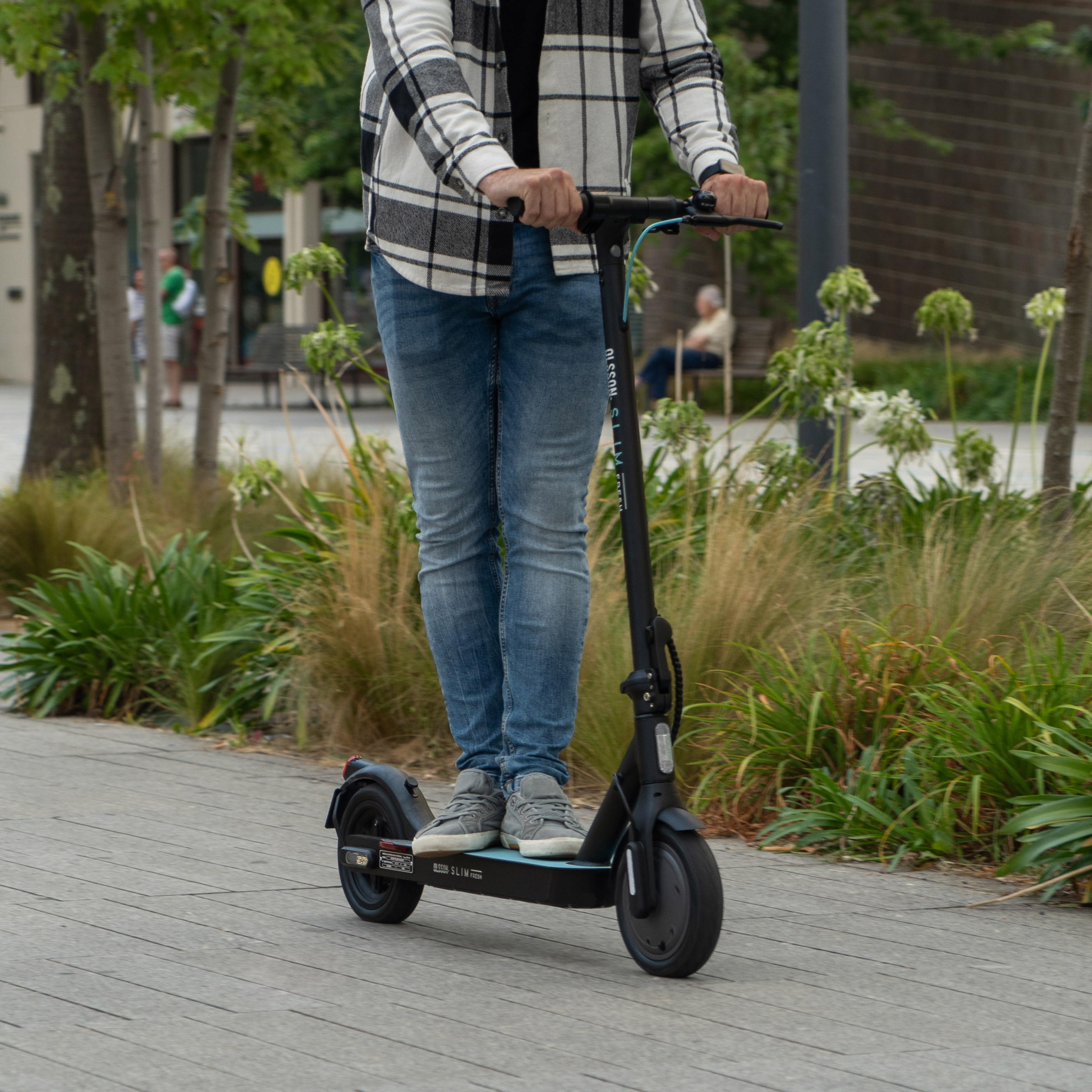 Persona montando un patinete eléctrico negro en un camino pavimentado en un parque, árboles atrás.