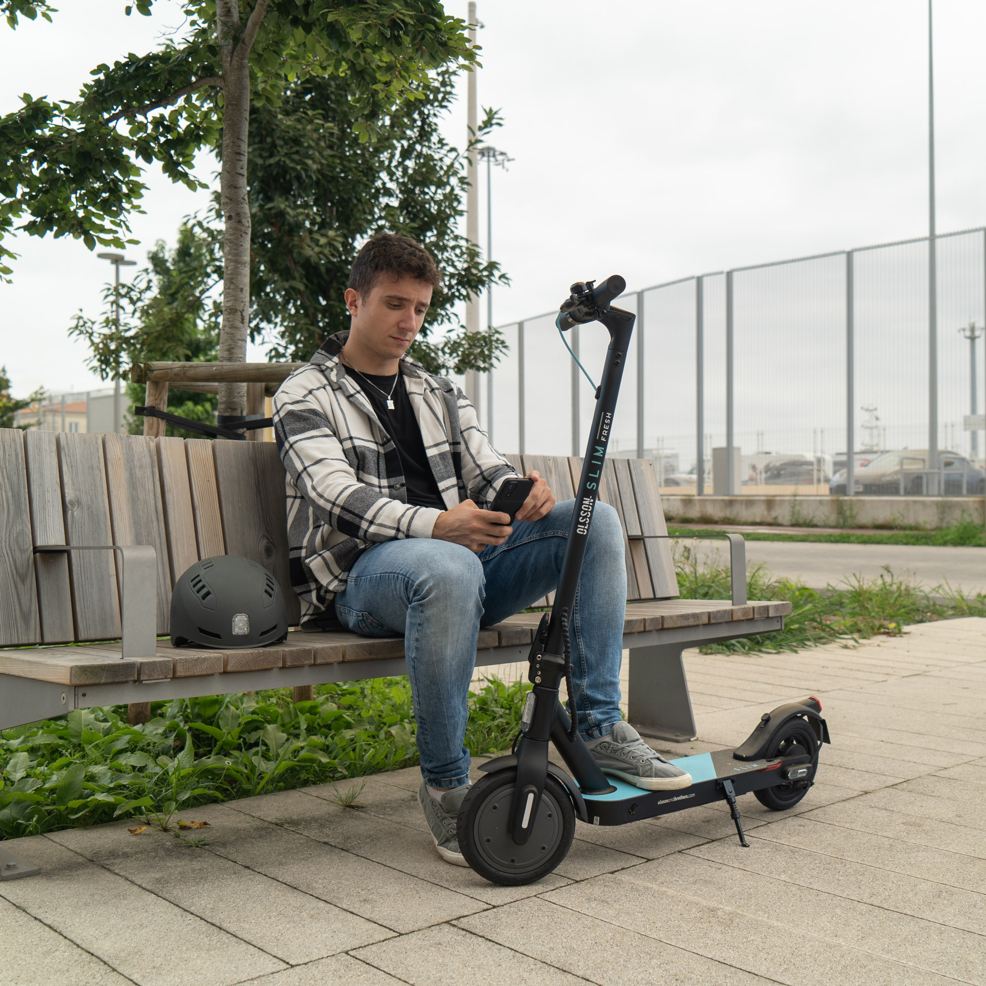 Hombre sentado en un banco con un scooter eléctrico, revisando su teléfono, con un casco cerca.