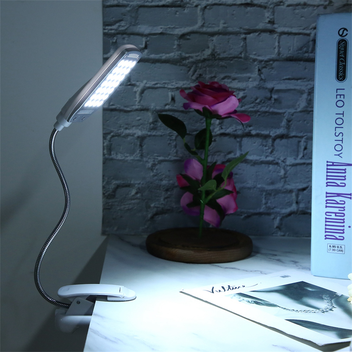 Eine flexible Schreibtischlampe beleuchtet einen Schreibtisch mit einem Buch, einer Rose und einer Backsteinmauer.