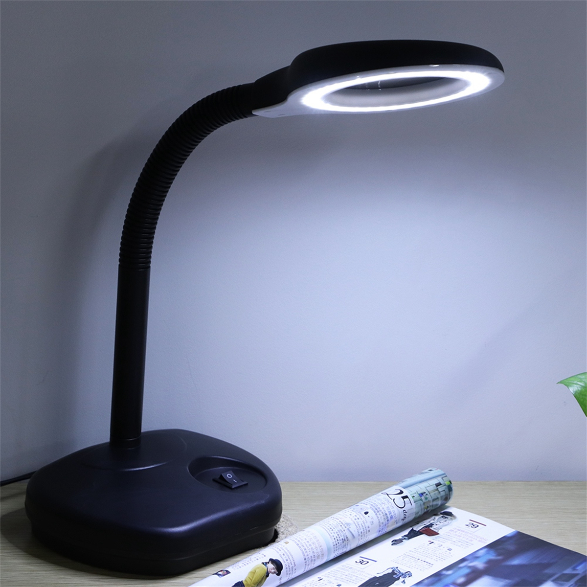 Eine schwarze Schreibtischlampe mit Lupenkopf, beleuchtet, auf einem Schreibtisch mit einer Zeitschrift.
