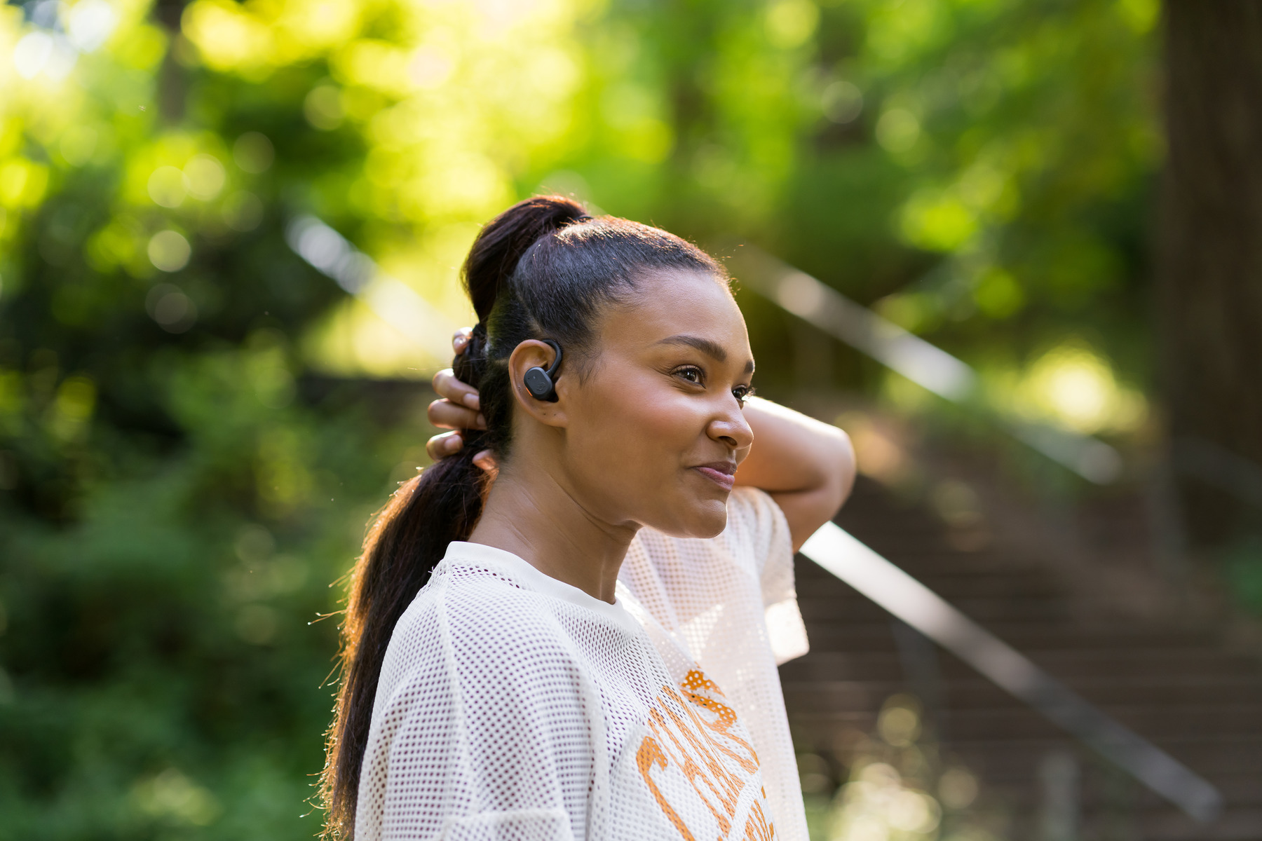Woman with ponytail wearing wireless earbuds. Standing outdoors with blurred greenery in background.