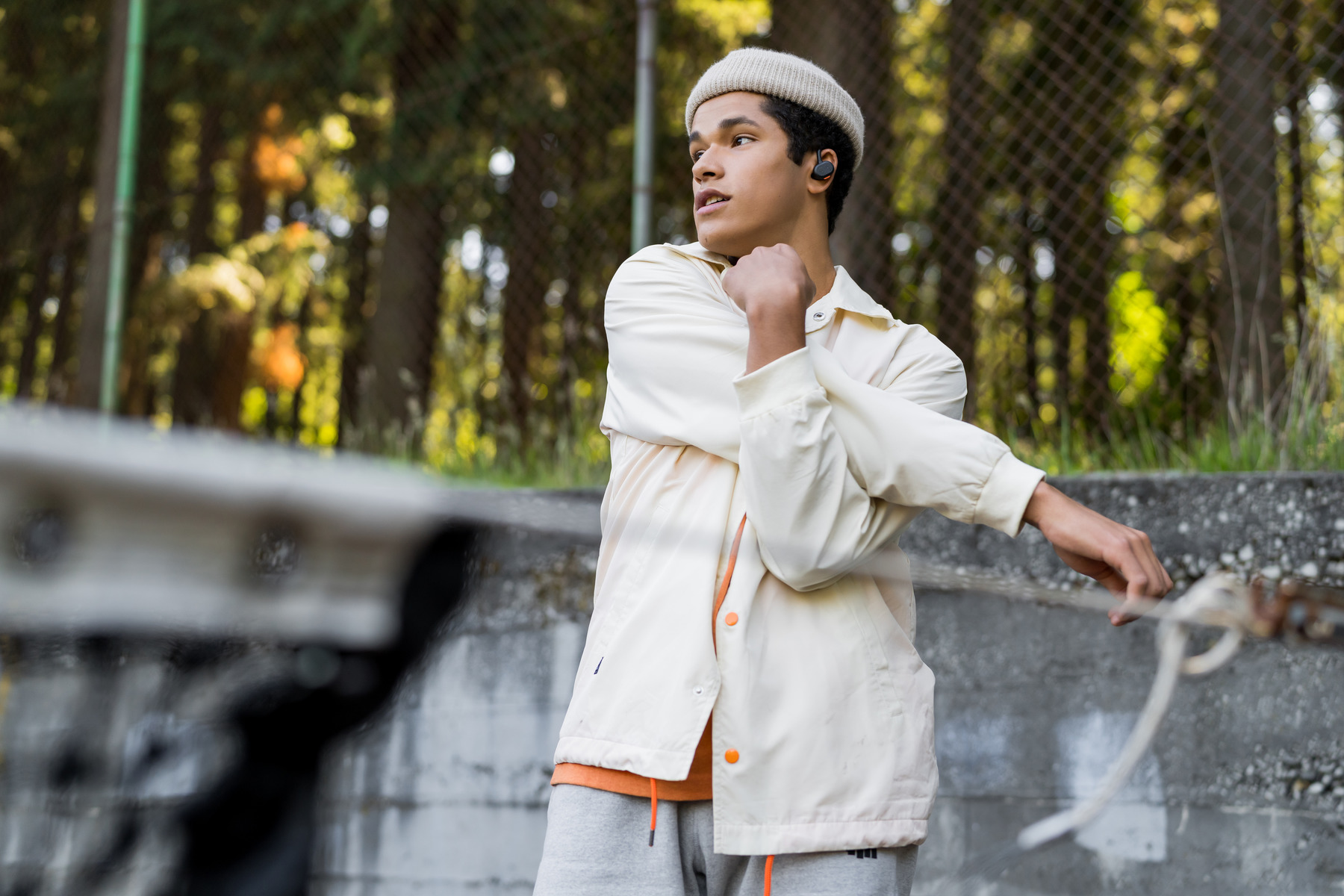 A young man in a beanie and jacket stretches outdoors, with a green forest in the background.