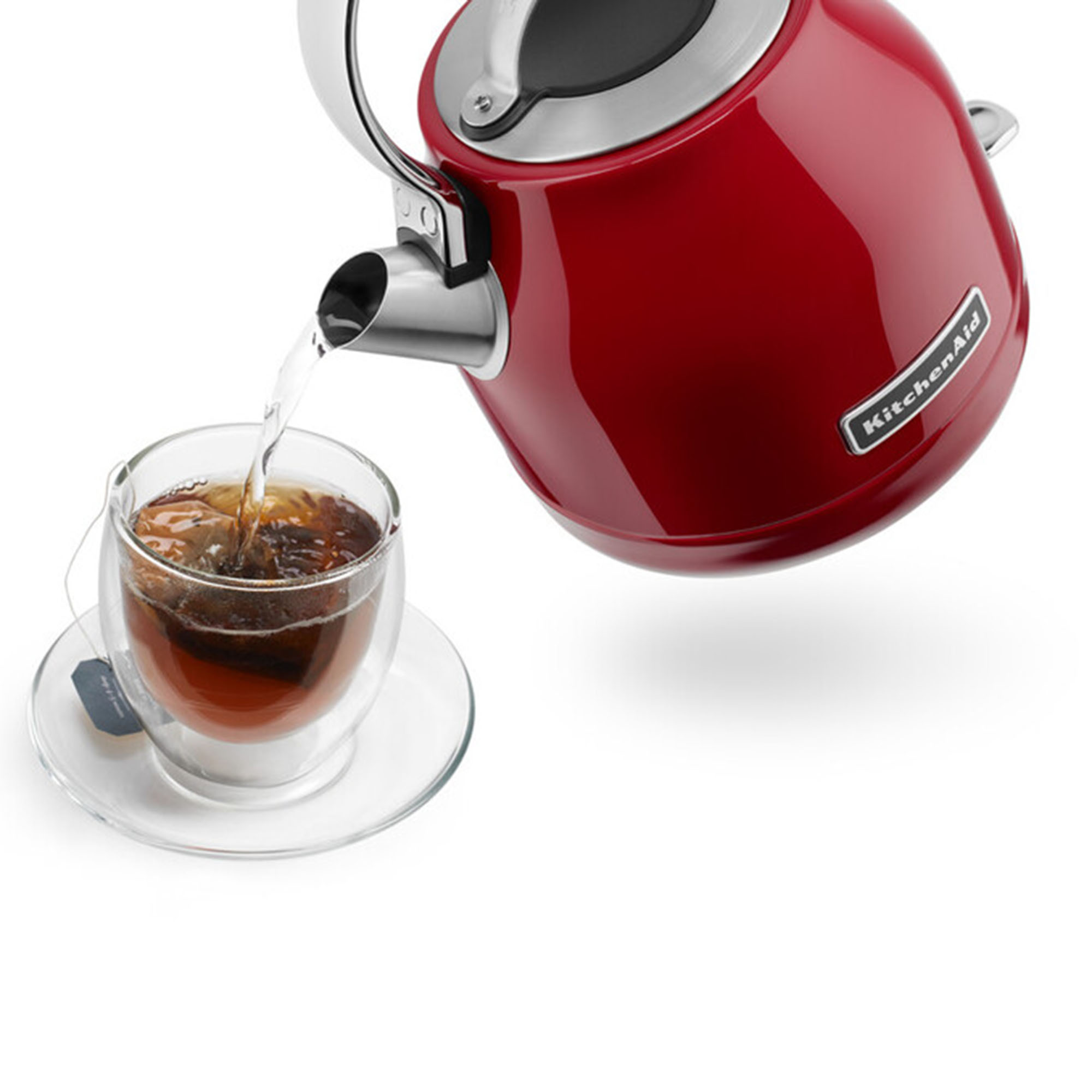 Hervidor KitchenAid rojo vertiendo agua en una taza de vidrio con té, sobre fondo blanco.