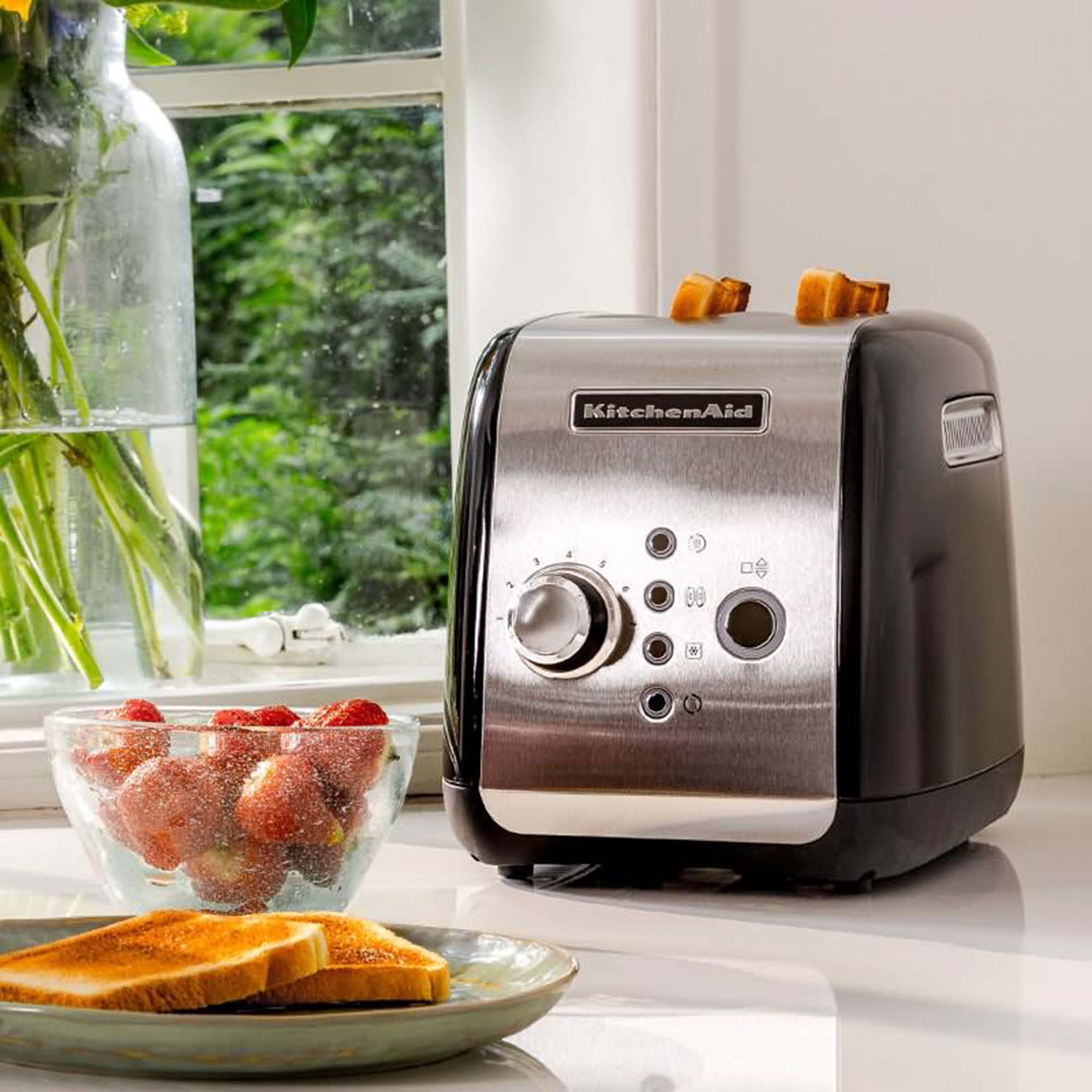 Black and silver KitchenAid toaster with bread and strawberries beside a window.