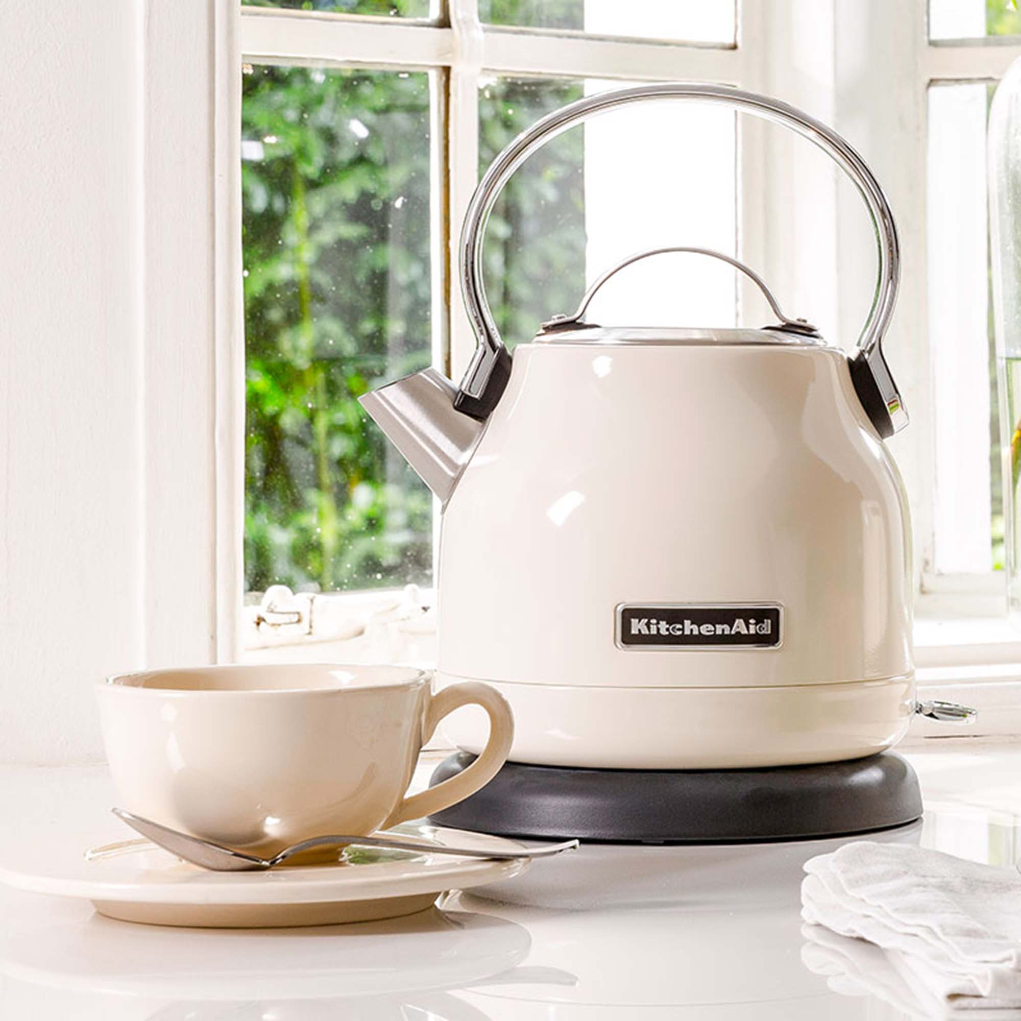 A cream-colored KitchenAid kettle and cup set on a white surface next to a window.