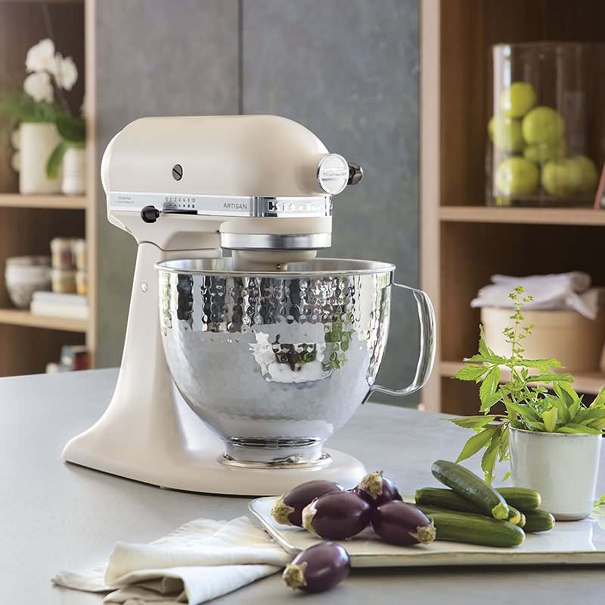 KitchenAid-Küchenmaschine, cremefarben, mit Edelstahlschüssel. Gemüse und Dekoration im Hintergrund.