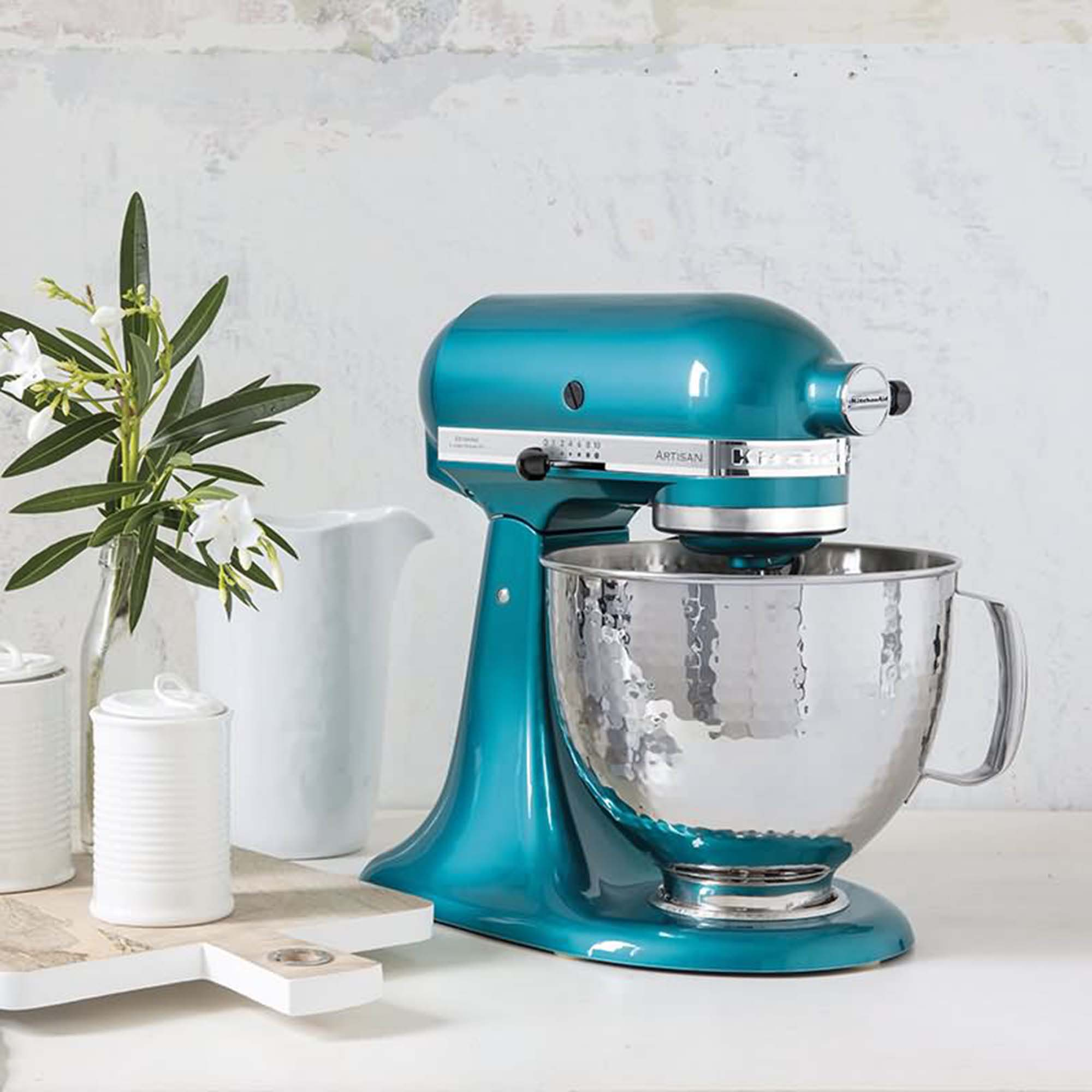 Ein türkisfarbener KitchenAid-Küchenmixer mit einer Edelstahlschüssel auf einer weißen Oberfläche mit Blumen und einem Krug.