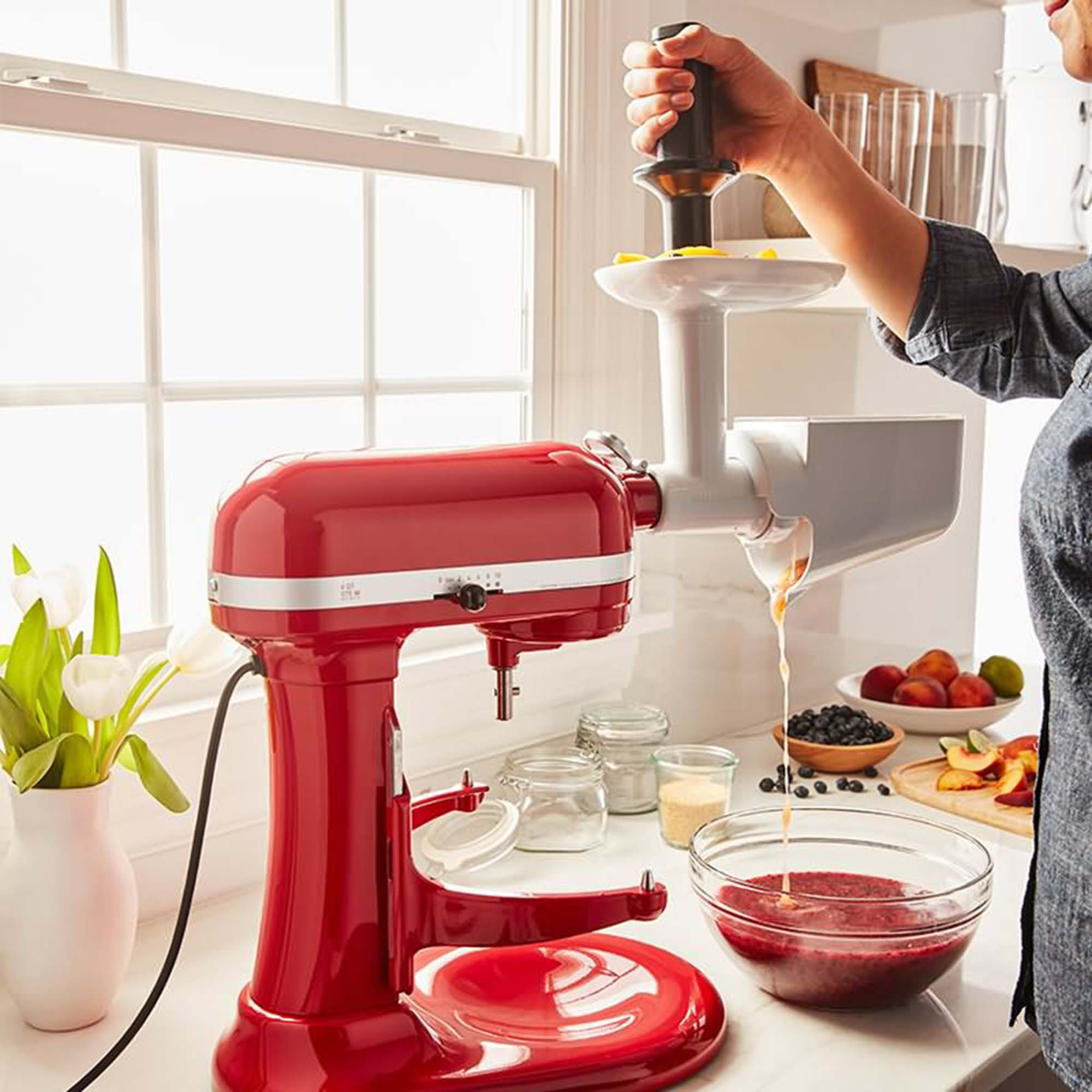 Un mixer da cucina rosso con accessorio spremiagrumi, una persona aggiunge frutta. Ingredienti e ciotole visibili.