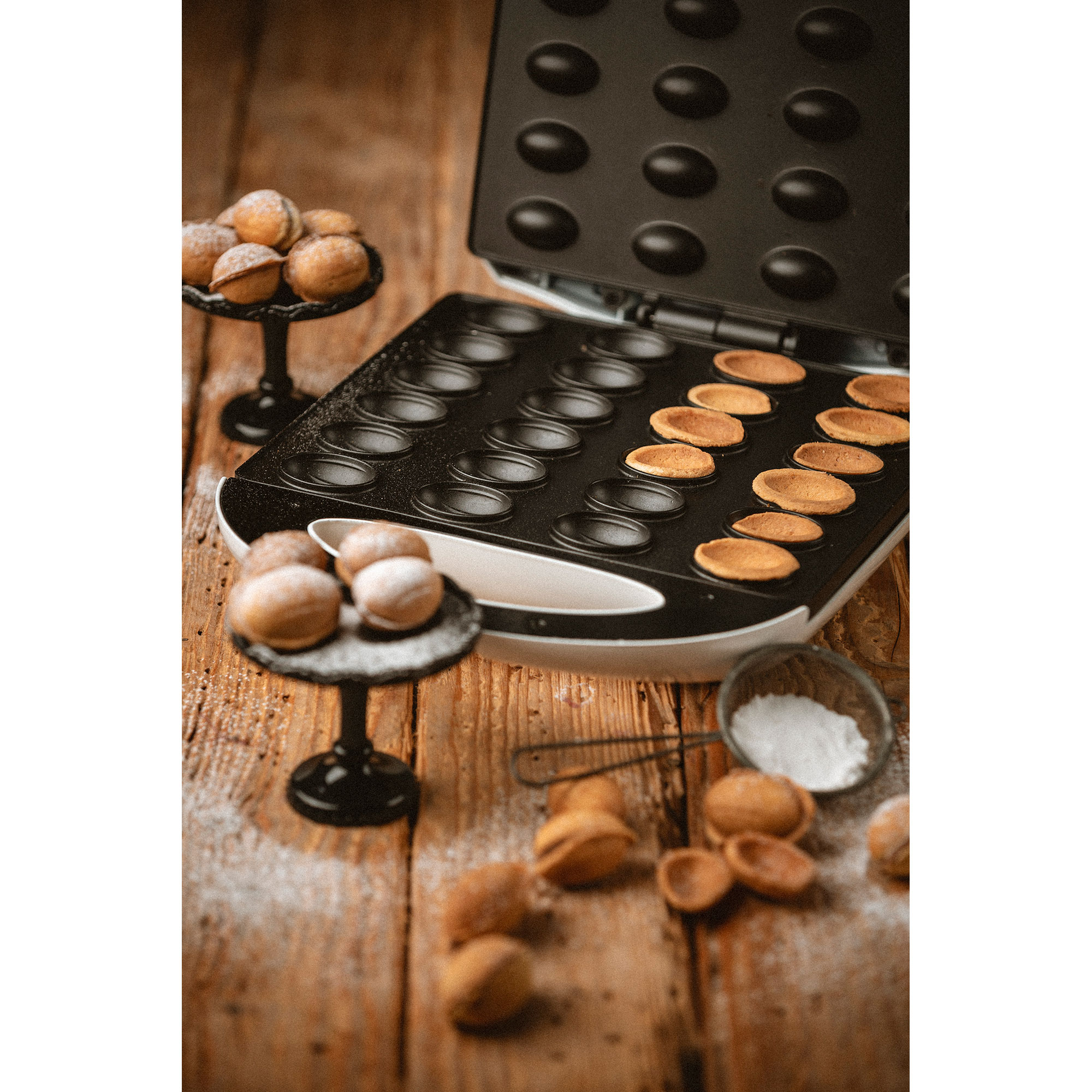 Otwarta gofrownica z upieczonymi goframi, obok talerzy z goframi posypanymi cukrem pudrem.