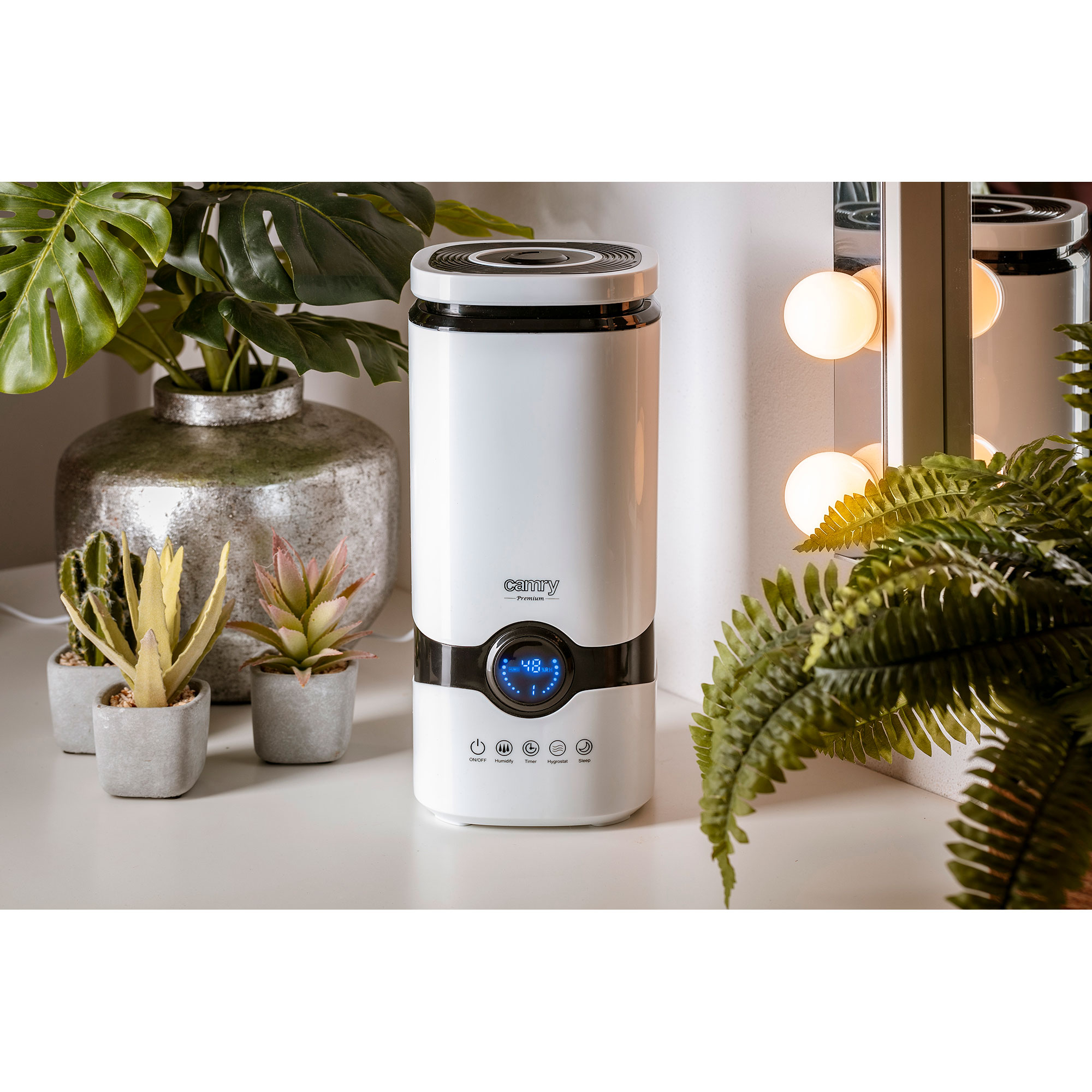 Biały nawilżacz z wyświetlaczem, otoczony roślinami i elementami dekoracyjnymi na białej powierzchni.