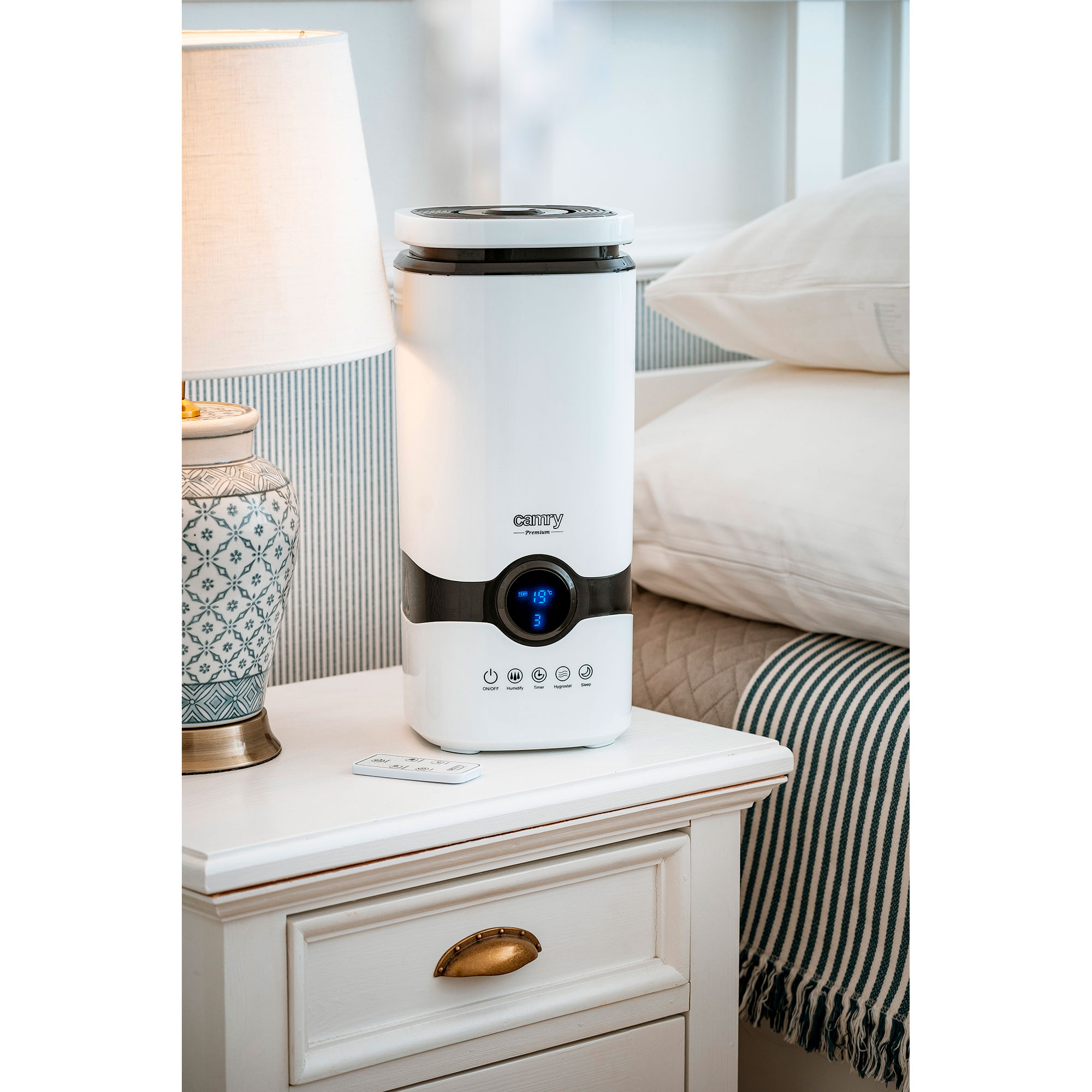 Biały nawilżacz powietrza na białej szafce nocnej obok łóżka, z pilotem.