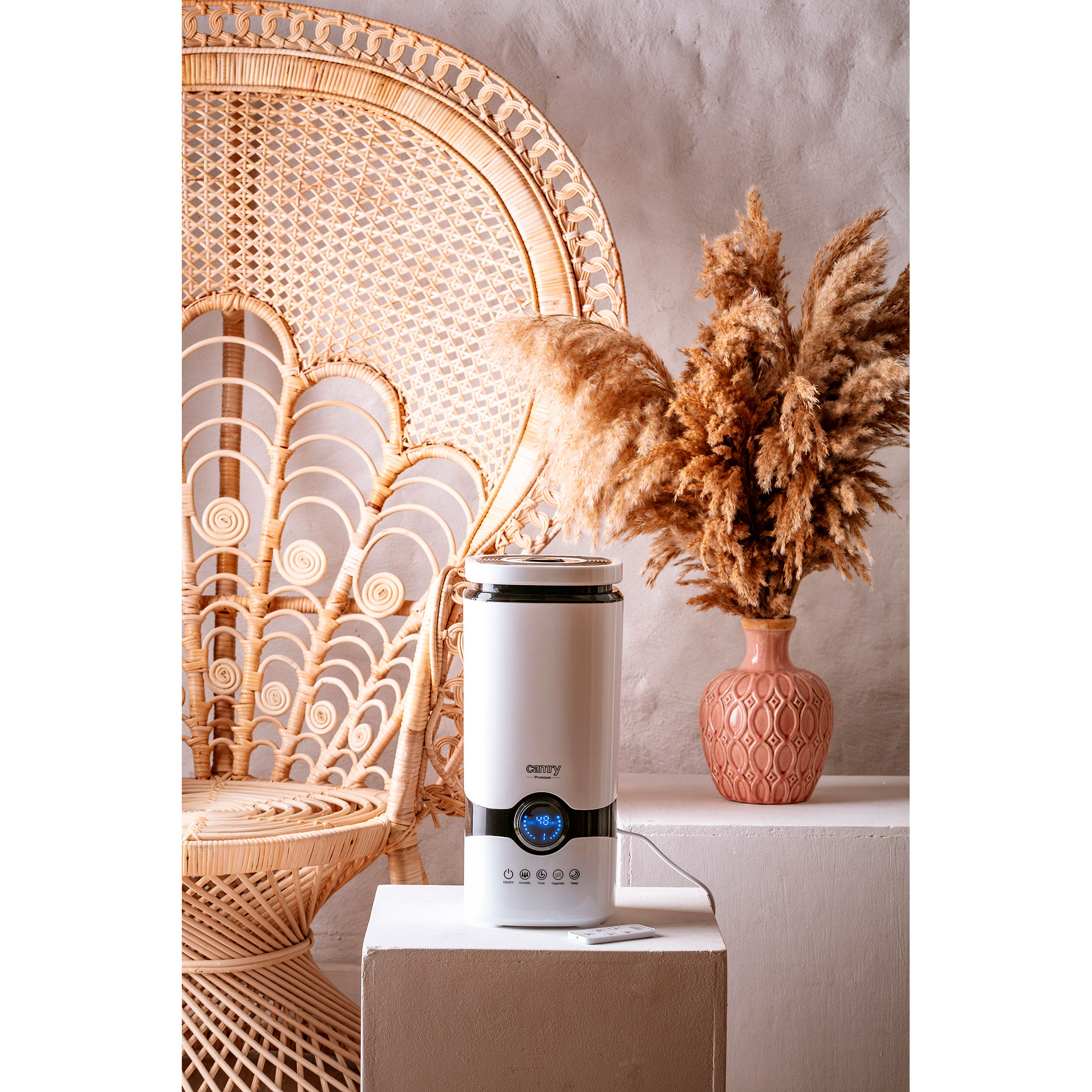Biały nawilżacz powietrza z cyfrowym wyświetlaczem, obok wiklinowego krzesła i suchych kwiatów.