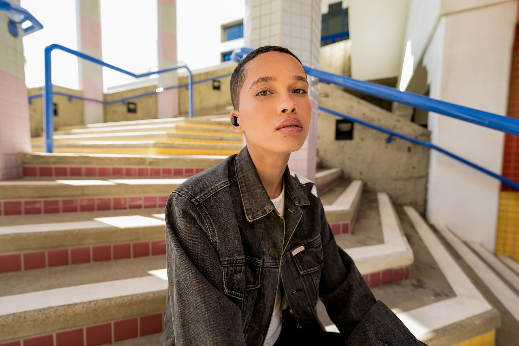Kobieta siedząca na schodach, w dżinsowej kurtce, ze słuchawkami, patrząca w kamerę.