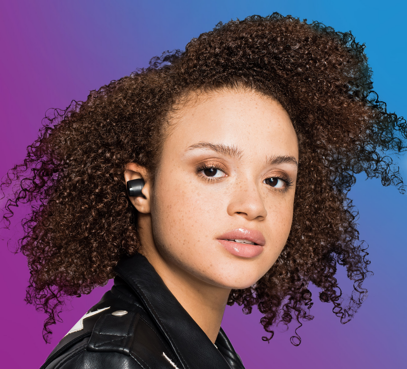 Eine Frau mit lockigem Haar trägt schwarze Ohrhörer und eine Lederjacke, mit blauem und lila Hintergrund.