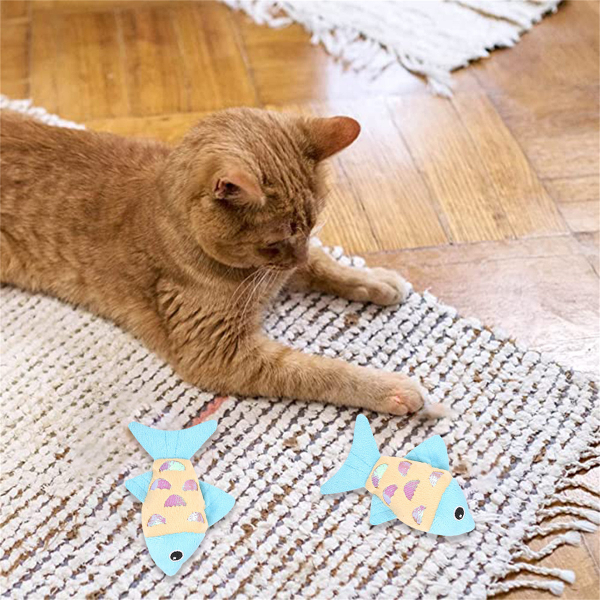Orange Katze spielt mit zwei blauen Fischspielzeugen auf einem gemusterten Teppich.