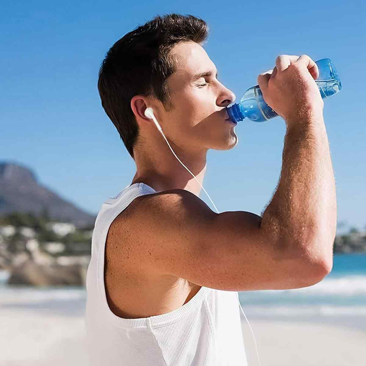 Mann trinkt Wasser, trägt Kopfhörer an einem sonnigen Strand mit Berg im Hintergrund.