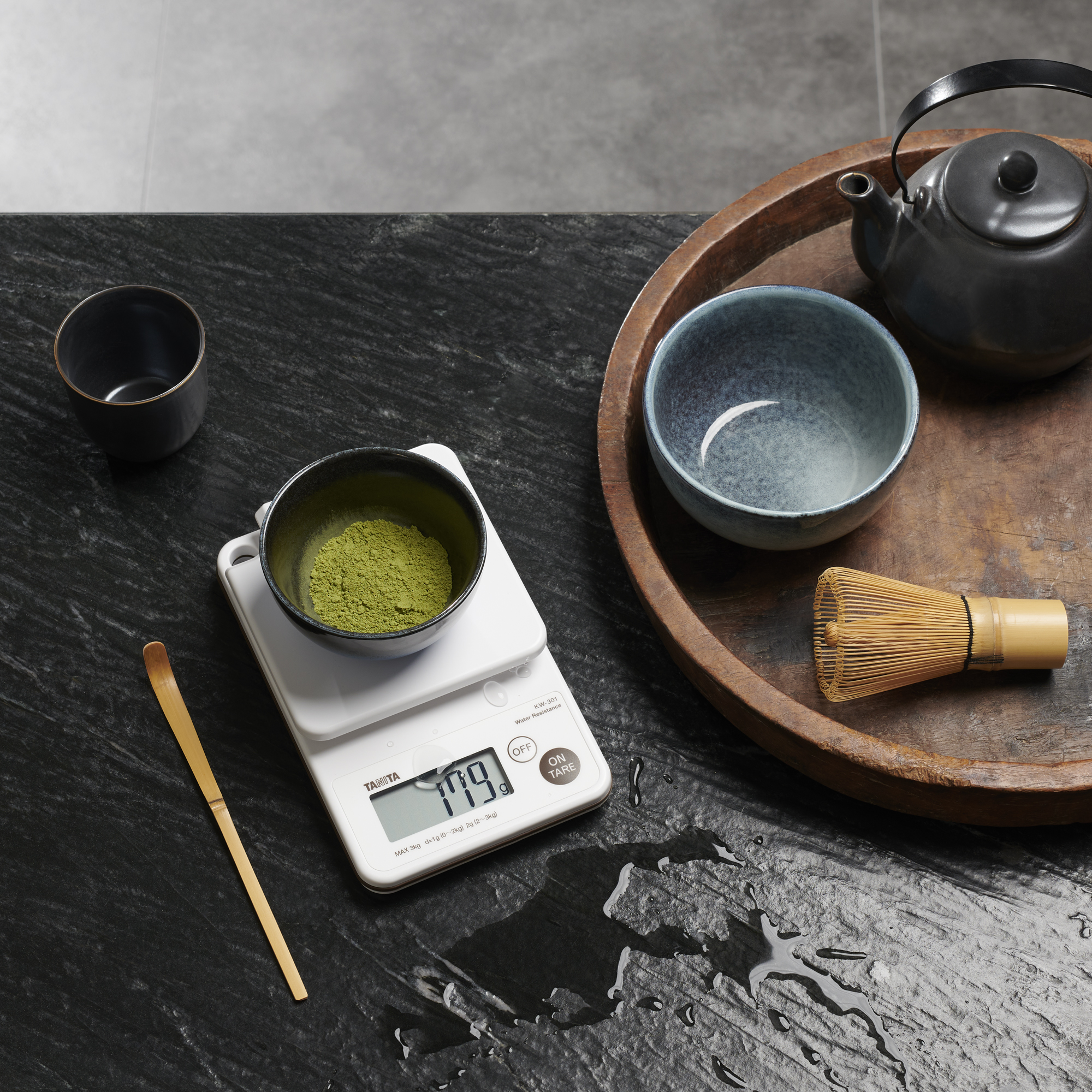 Matcha-theeset op een zwart oppervlak. Er is een kom met matcha, een weegschaal en een garde aanwezig.