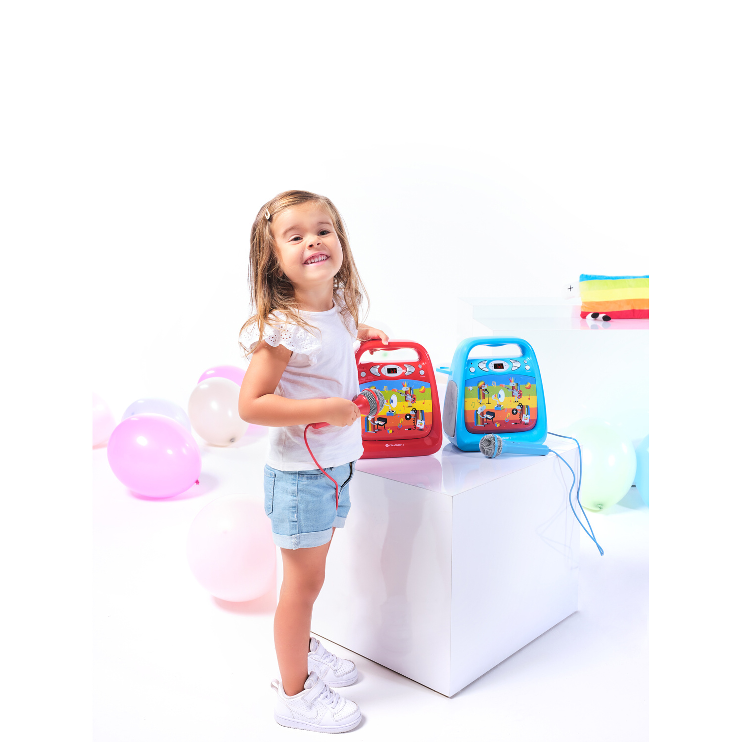 Ein kleines Mädchen steht neben zwei Spielzeug-Karaoke-Geräten und Ballons.