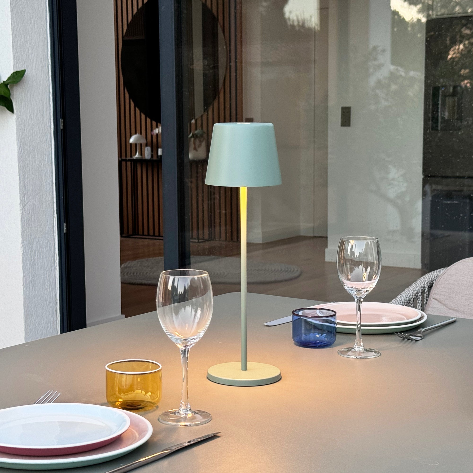 Eine kleine grüne Lampe auf einem Tisch mit zwei Gläsern, Tellern und einem blauen Behälter. Umgebung im Freien.