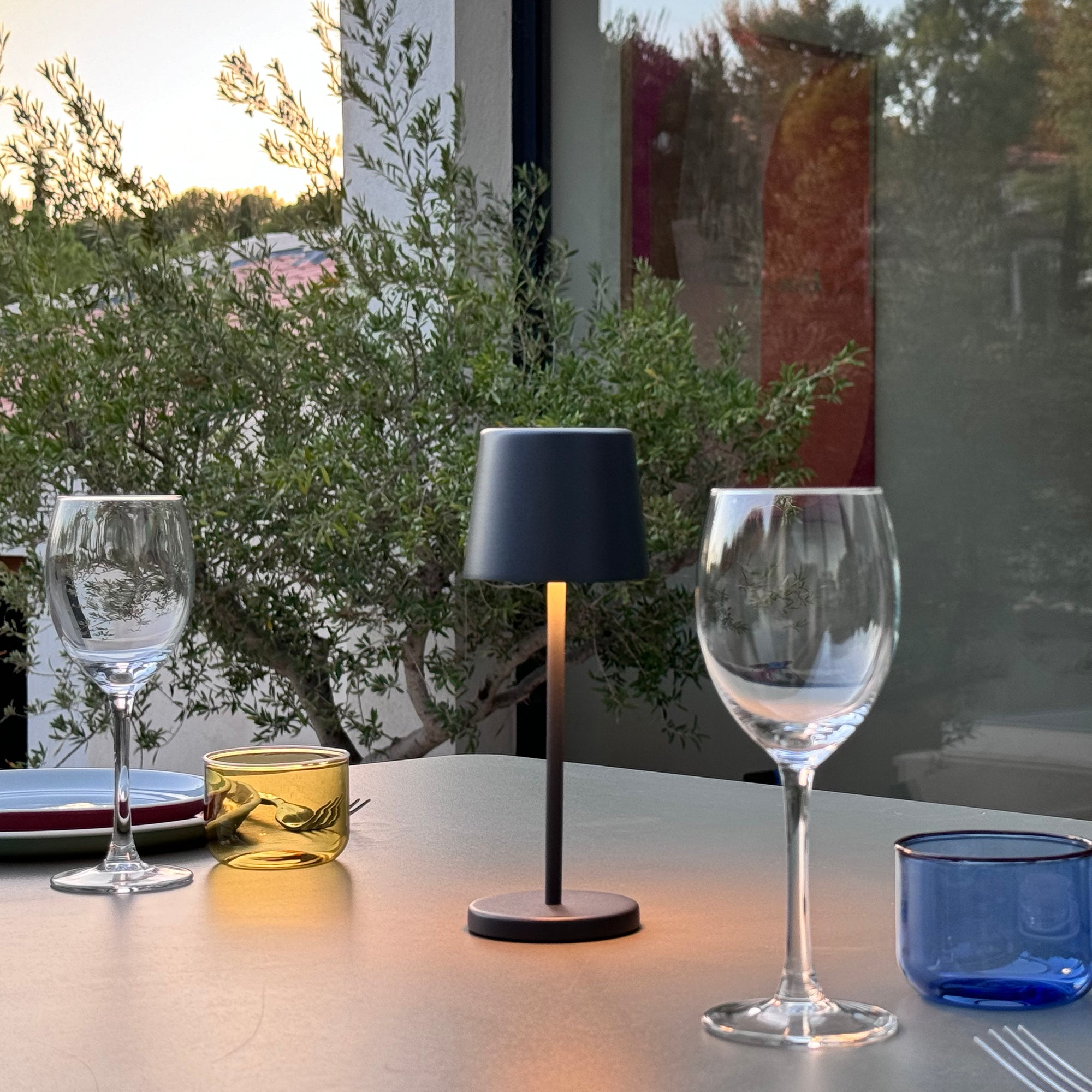 Tisch im Freien mit Gläsern, einer Lampe und Grün im Hintergrund.