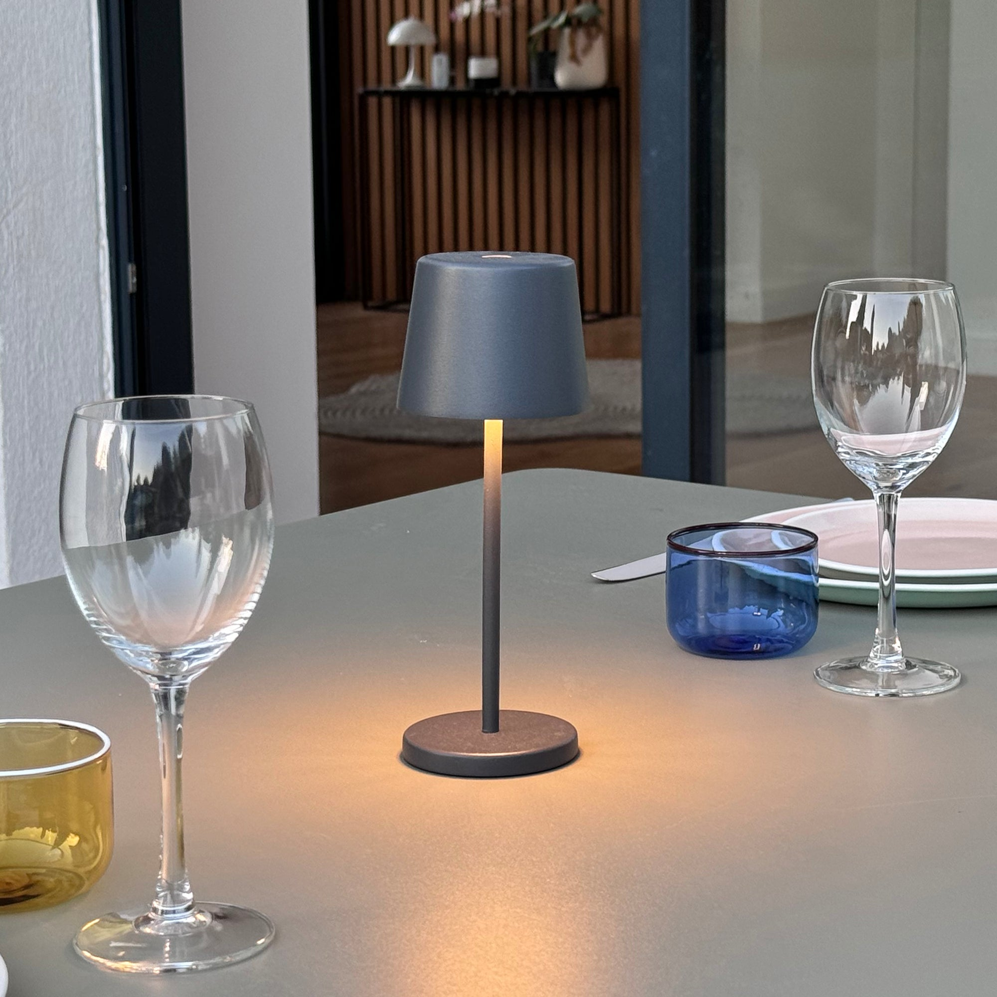 Tisch mit Lampe, Gläsern und blauer Schale. Hintergrund zeigt Holzpaneel und Tür.